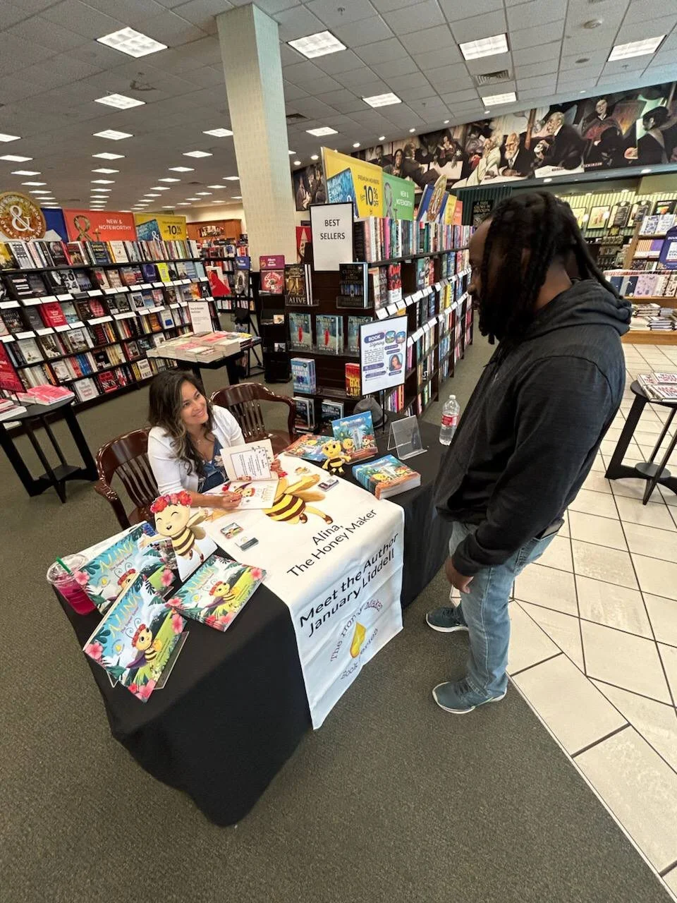 A woman sits behind a table with children's books and promotional materials at a book signing event in a bookstore, while a man stands in front of her, looking at the books.
