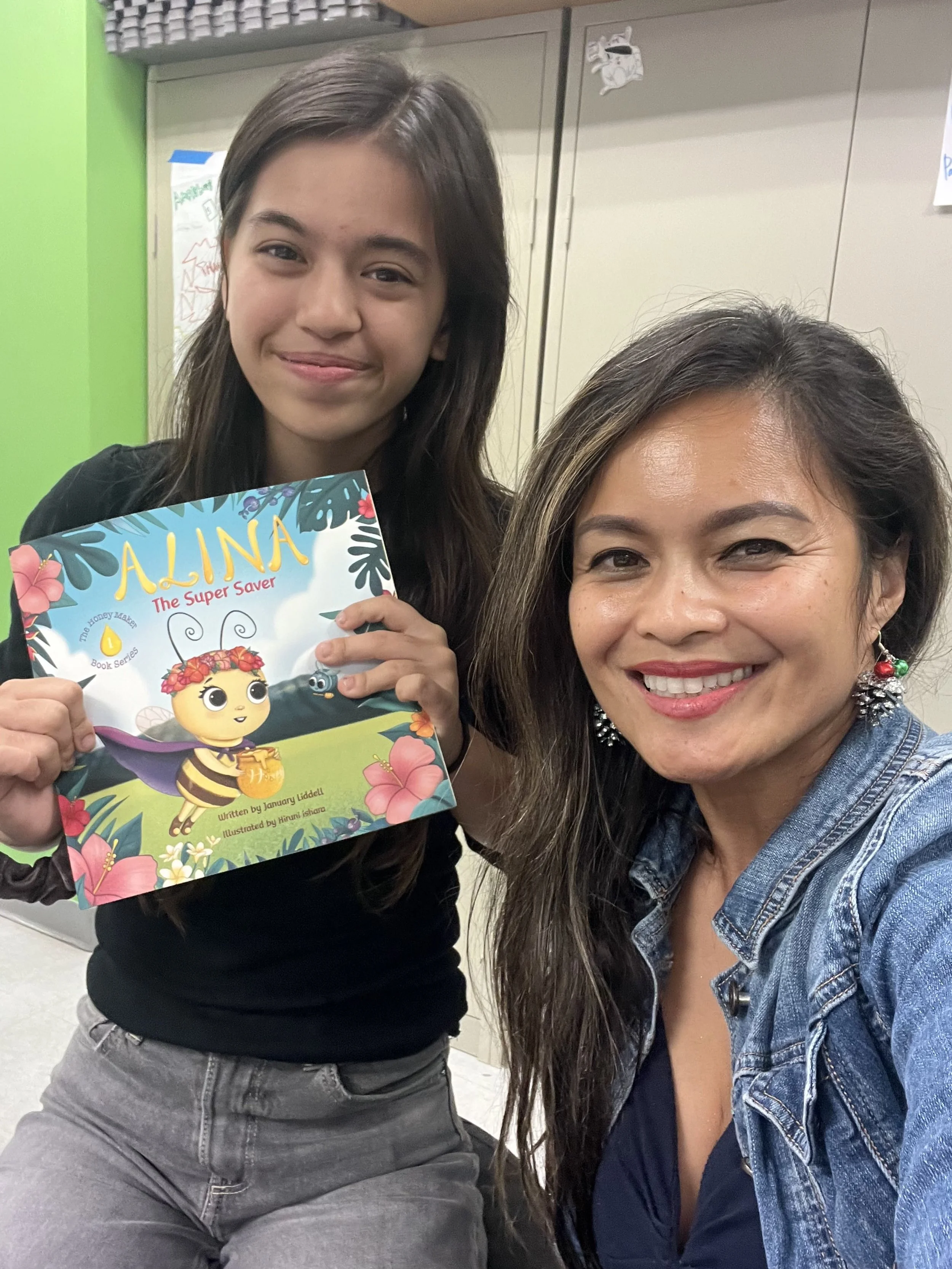 A young girl holding a colorful children's book titled 'Alina The Super Saver' with an illustrated bee character, next to a smiling woman with long dark hair wearing a denim jacket.