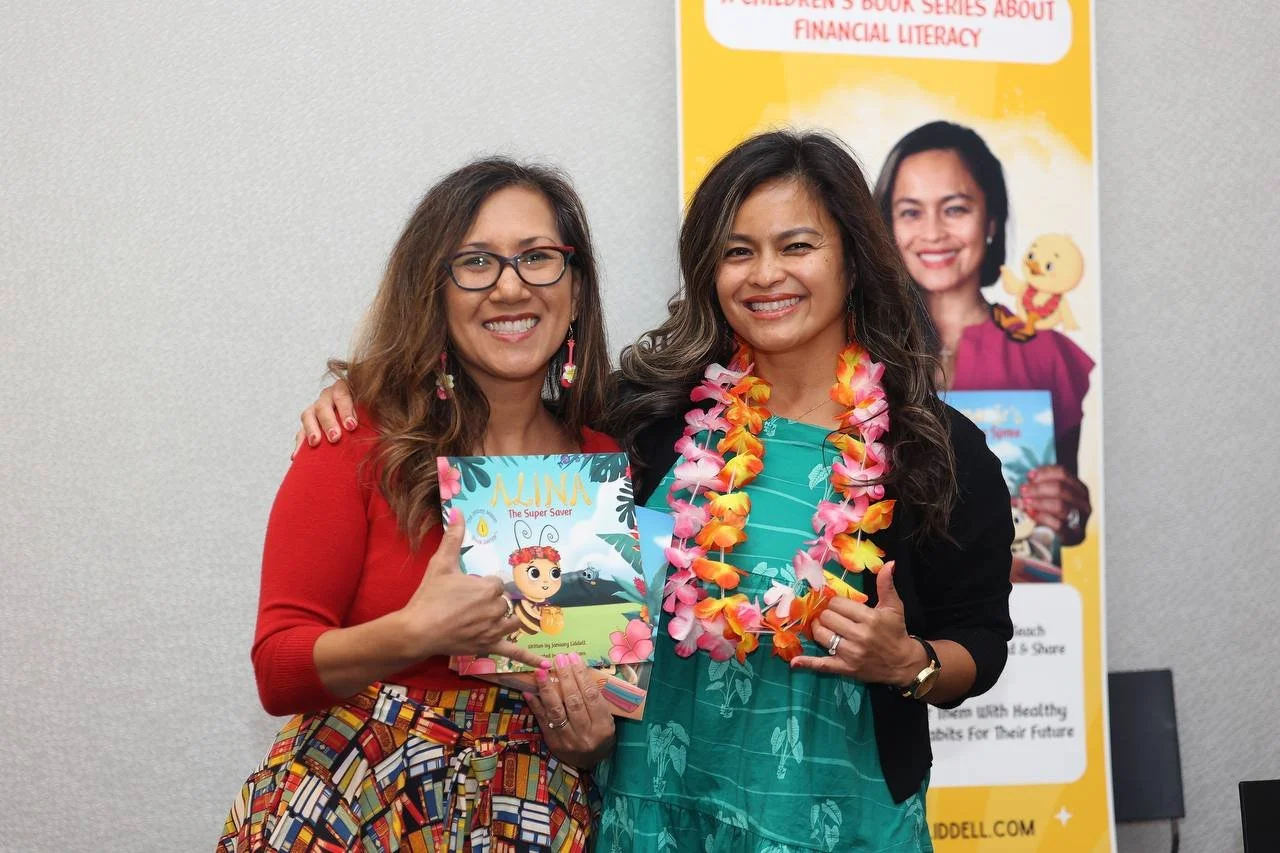 Two women smiling and posing together, one holding a children's book titled 'Alina The Super Saver'. The woman on the left is wearing glasses and a red top, while the woman on the right is wearing a turquoise dress and a colorful flower lei. Behind t