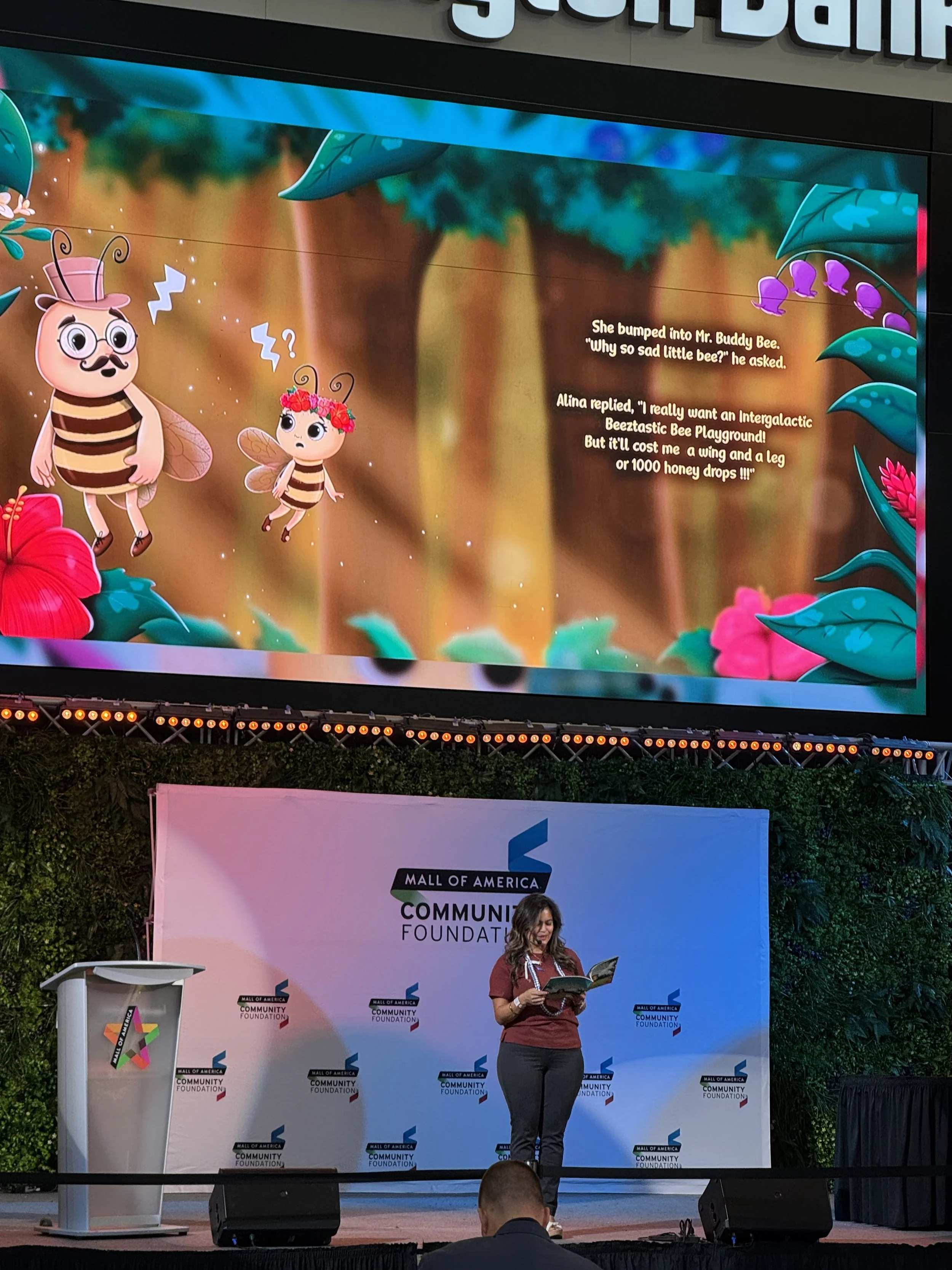 A woman is standing on a stage reading a book, with a large screen behind her displaying a colorful children's story about bees. The stage backdrop features the logos of the Mall of America Community Foundation, and there is a podium to her left.