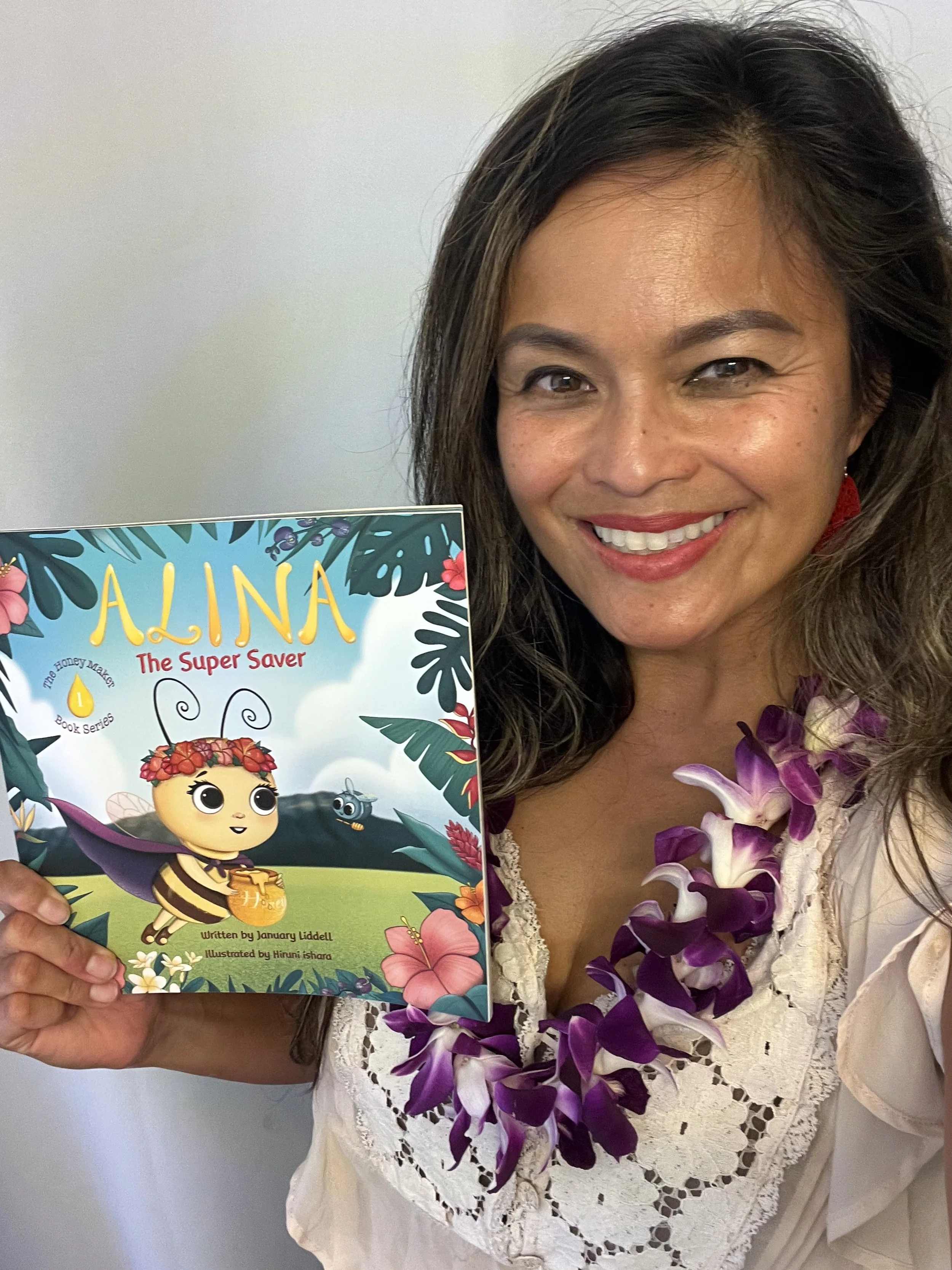 A woman with dark hair, wearing a white blouse with lace detailing and a purple and white flower lei, smiling and holding a colorful children's book titled 'Alina The Super Saver' featuring a cartoon bee character on the cover.