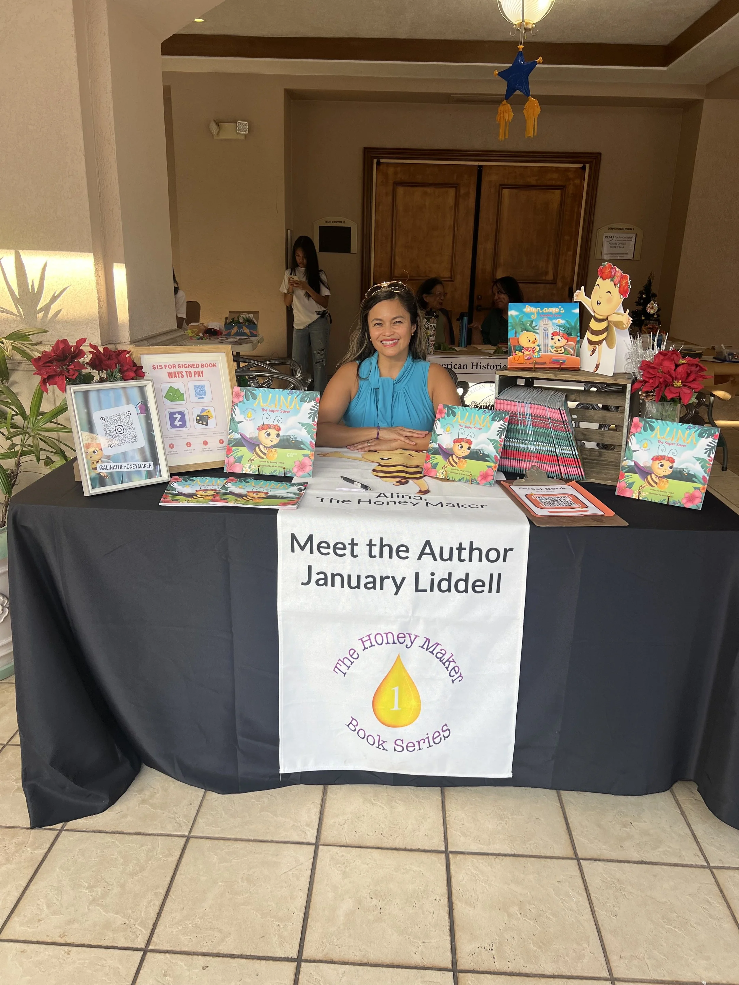 A woman sitting at a book signing table with a sign that reads "Meet the Author January Liddell" and "The Honey Maker Book Series." The table displays books, promotional materials, and a QR code. Behind her are other people and a small decorative sea