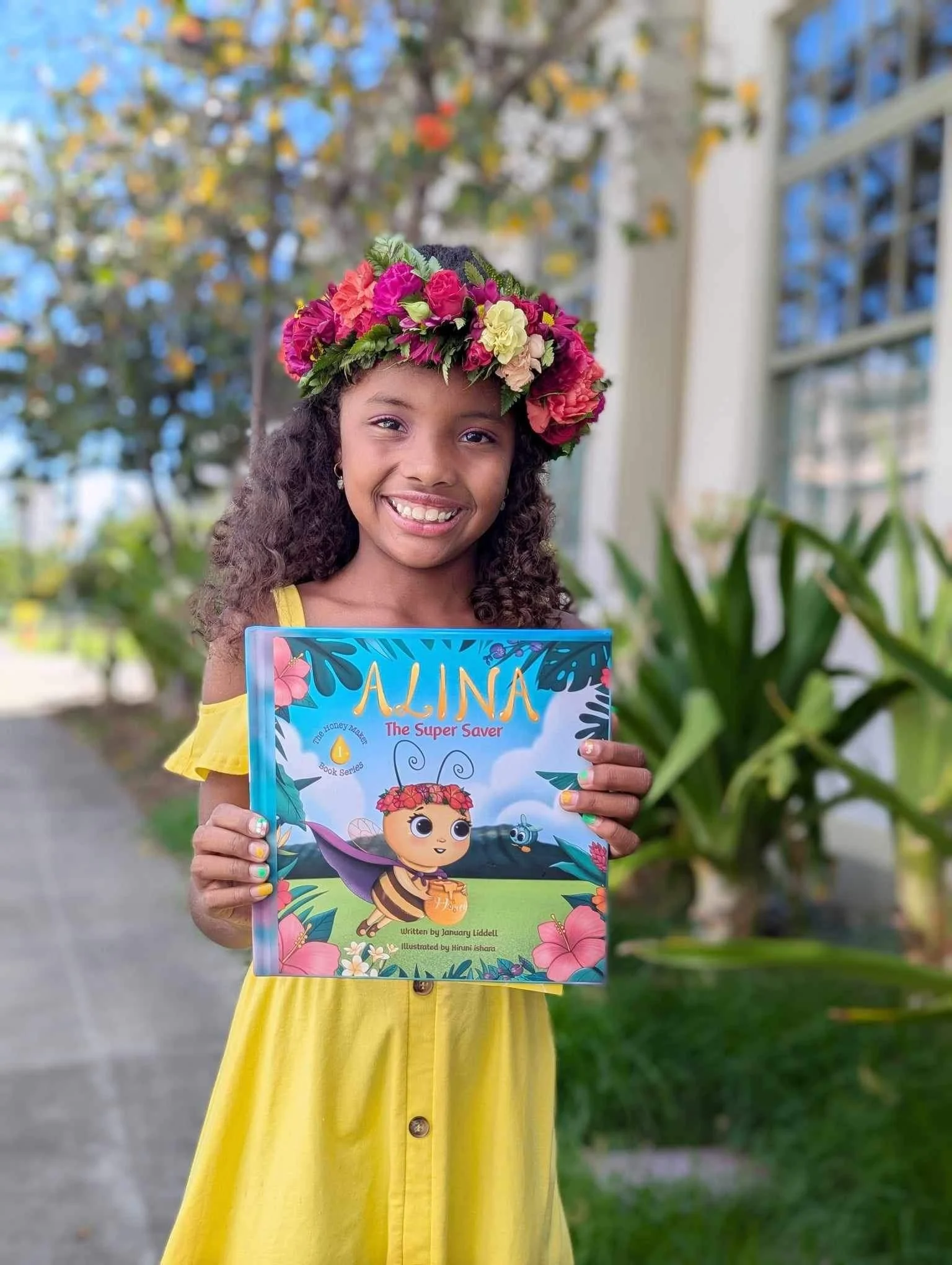 Smiling girl wearing a flower crown and a yellow dress, holding a colorful children's book titled 'Alina The Super Saver' outdoors with trees and a building in the background.