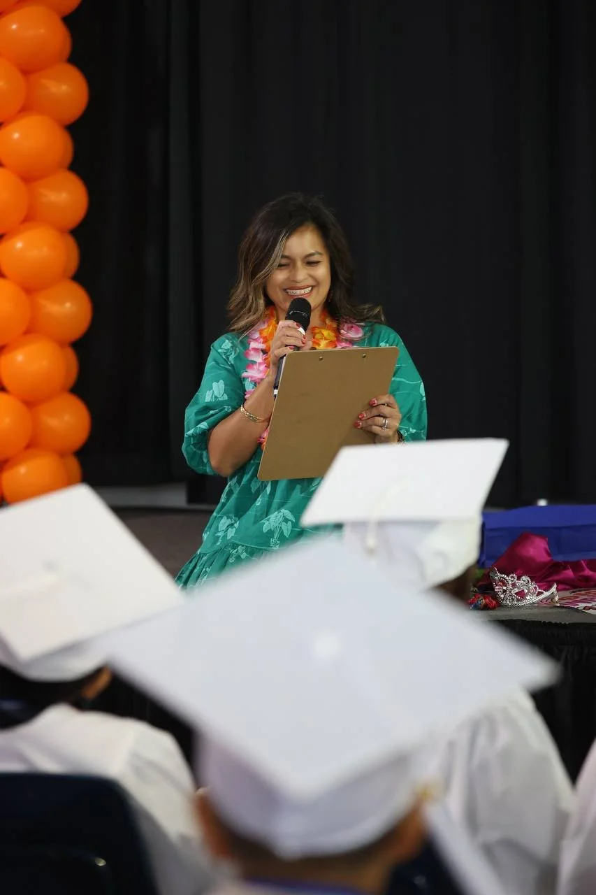 Woman at graduation ceremony speaking into a microphone, holding a clipboard, wearing a lei and a green dress, with orange balloons in the background.