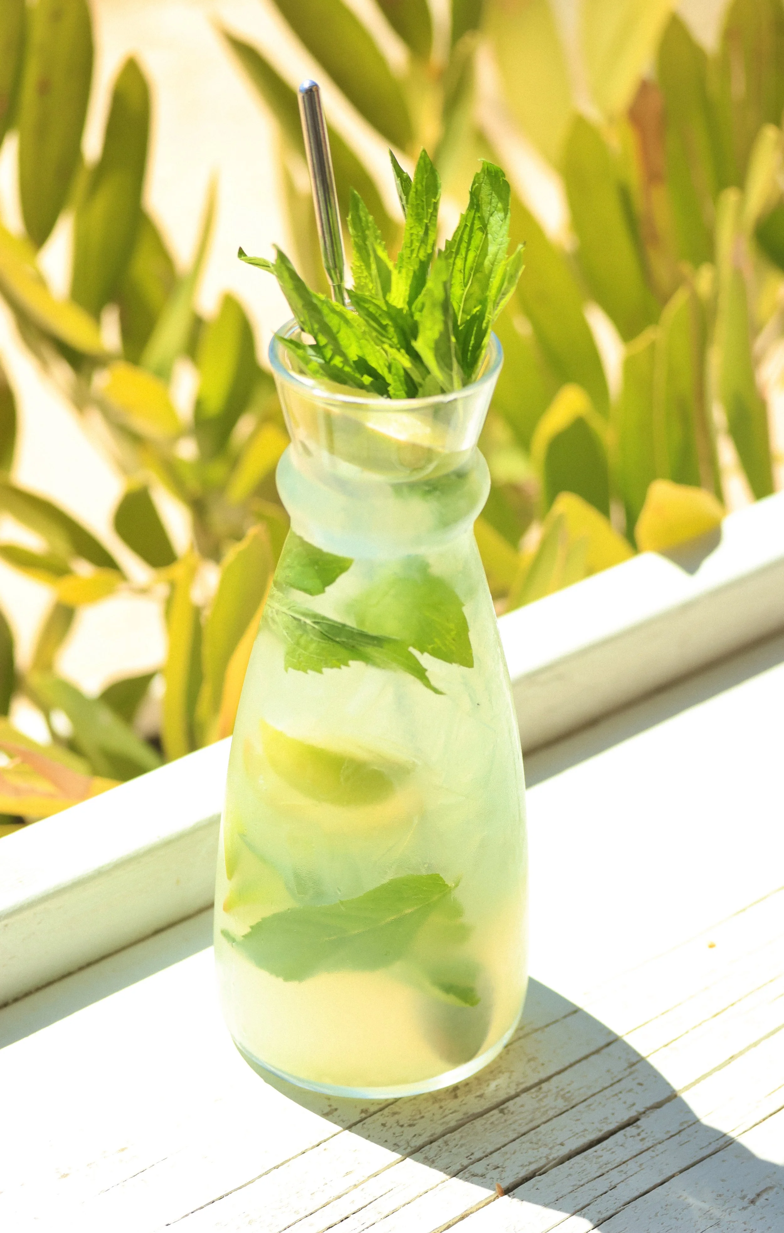 A glass bottle filled with lemonade garnished with fresh mint leaves and lemon slices, placed on a wooden surface outside with green plants in the background.
