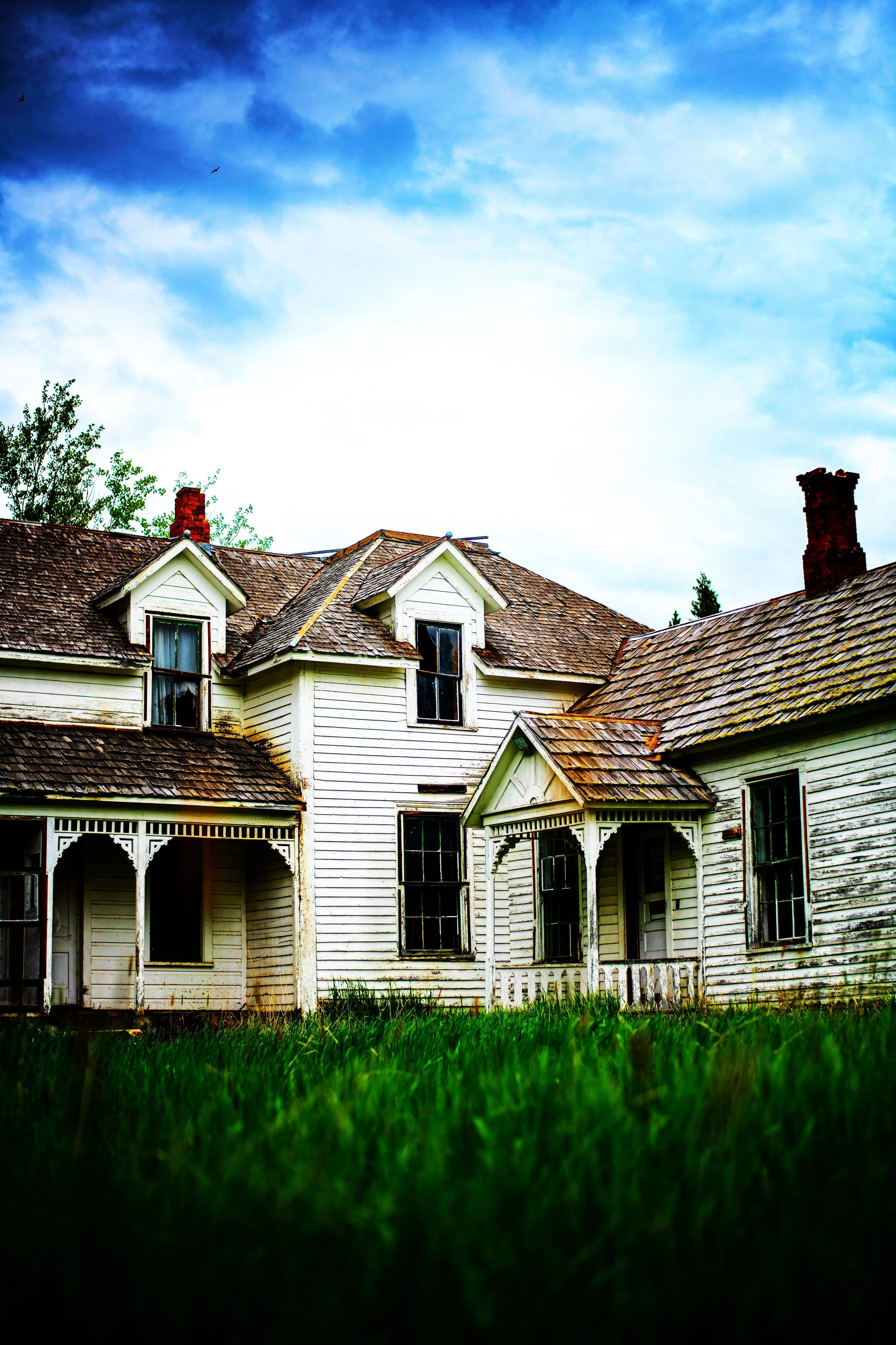 An old abandoned white wooden house with a slanted roof and multiple chimneys, surrounded by green grass and trees under a partly cloudy sky.