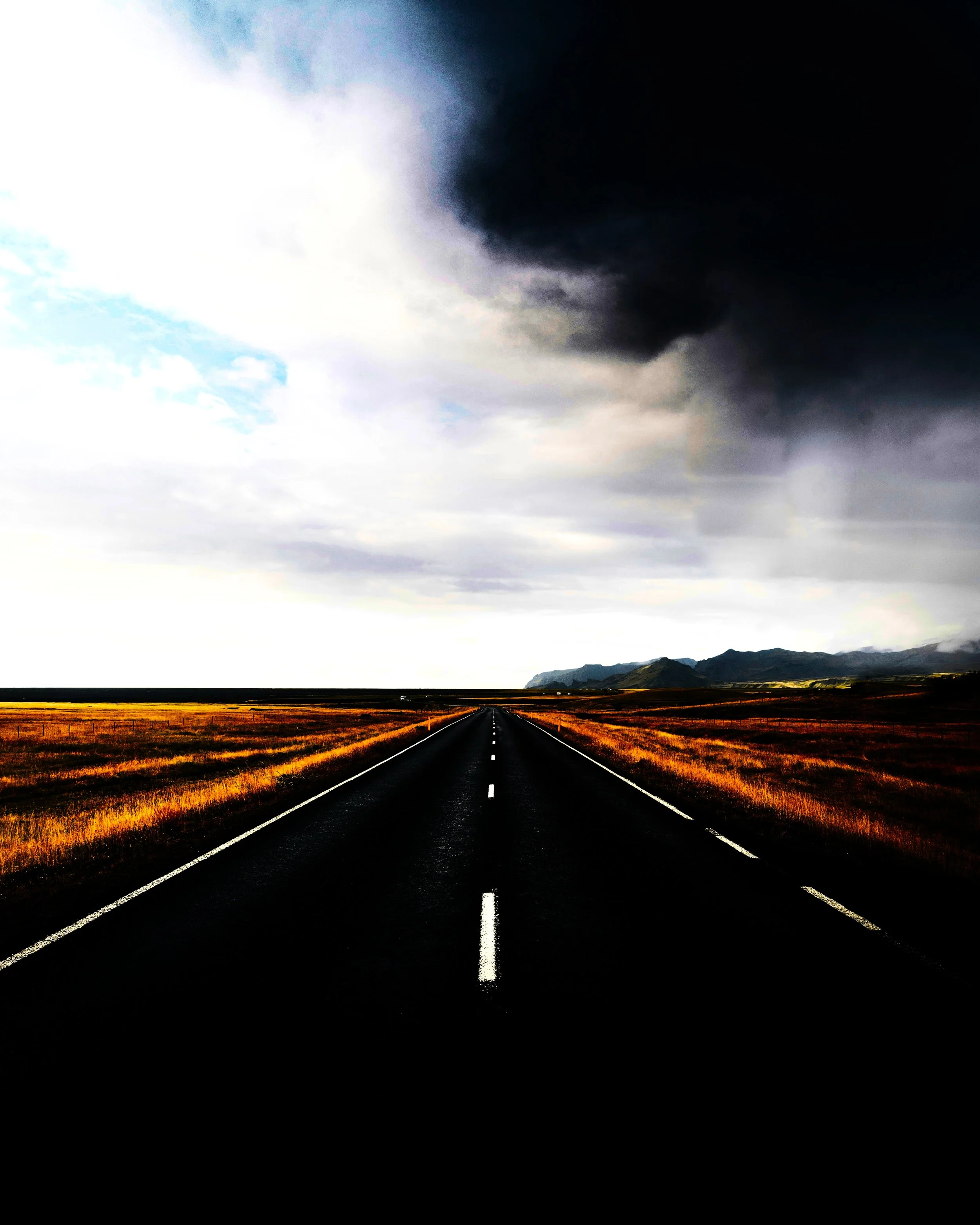 A long empty road stretching toward distant mountains under a dark, cloudy sky with patches of blue.