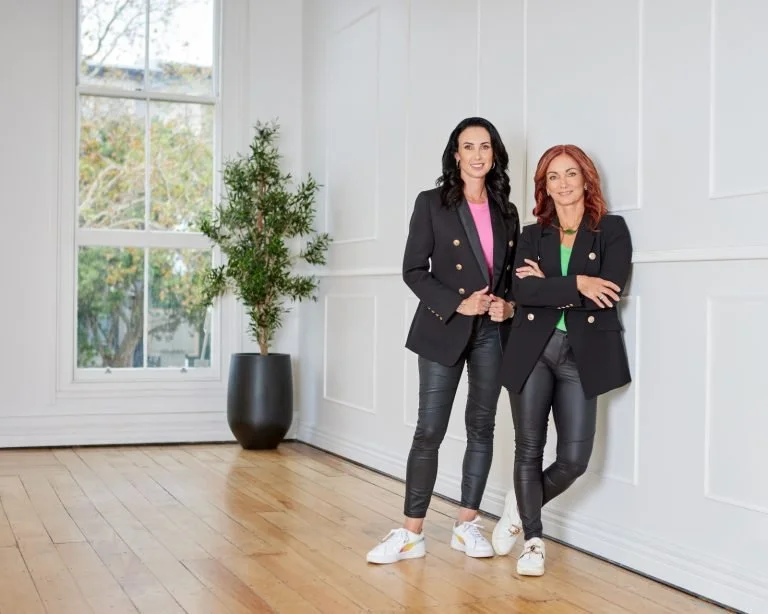Two women standing in a bright room with large window, white paneled walls, wooden floor, and a potted plant, dressed in black blazers, jeans, and sneakers.