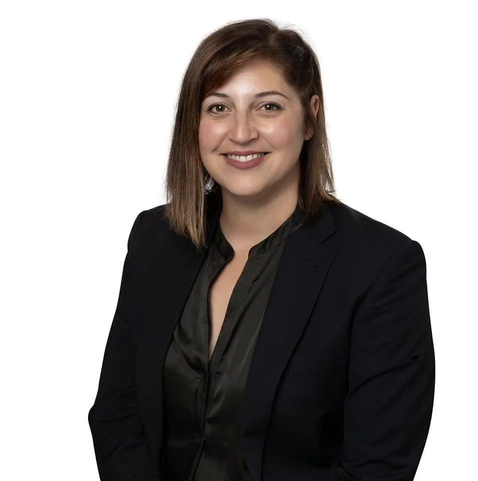Professional woman with shoulder-length brown hair, wearing a black blazer and black blouse, smiling against a plain white background.
