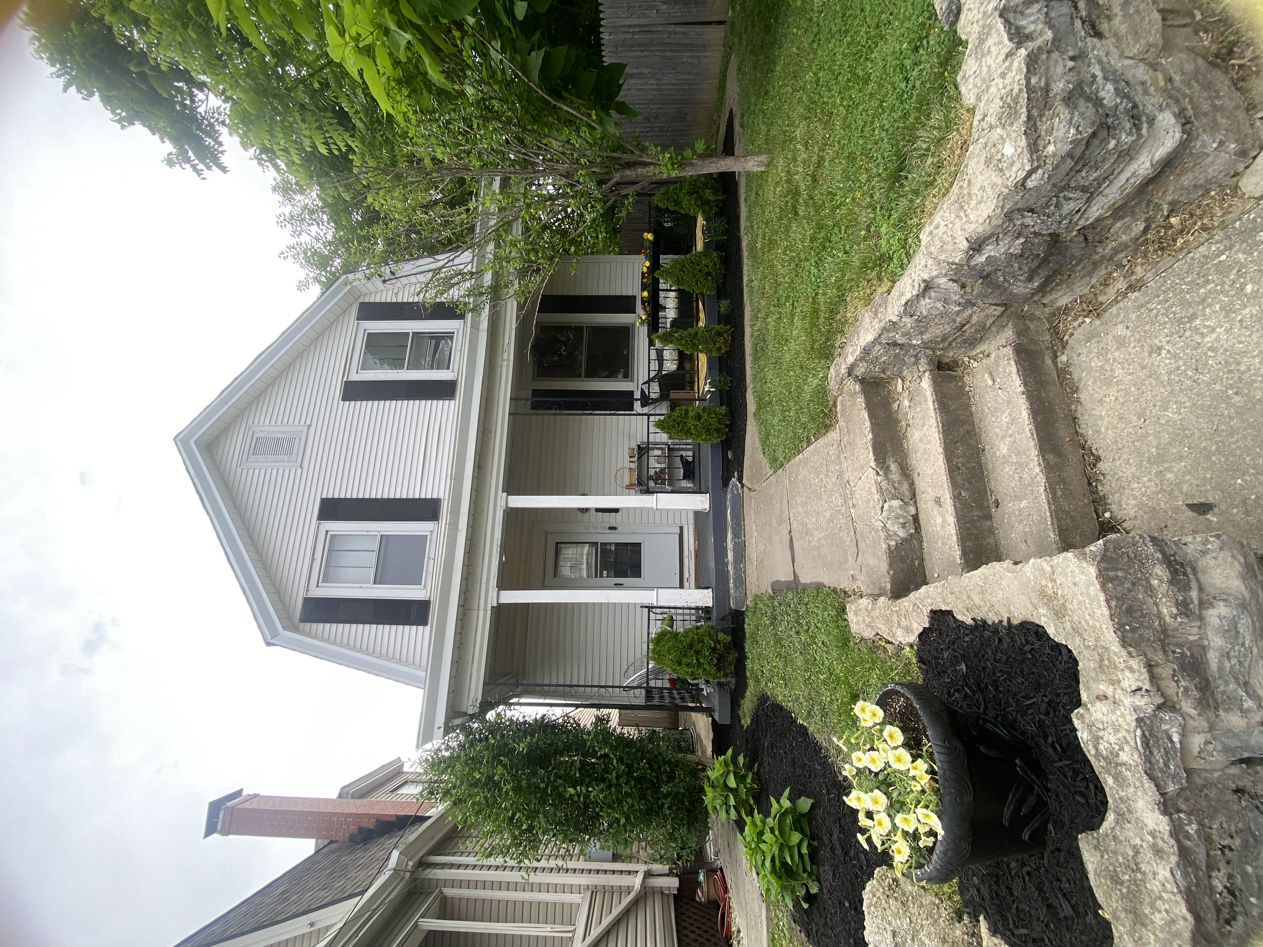 Front view of a white two-story house with black shutters, a porch with potted plants, and a landscaped yard with a small tree, bush, and flower bed with white and yellow flowers in a black pot surrounded by stone edging.