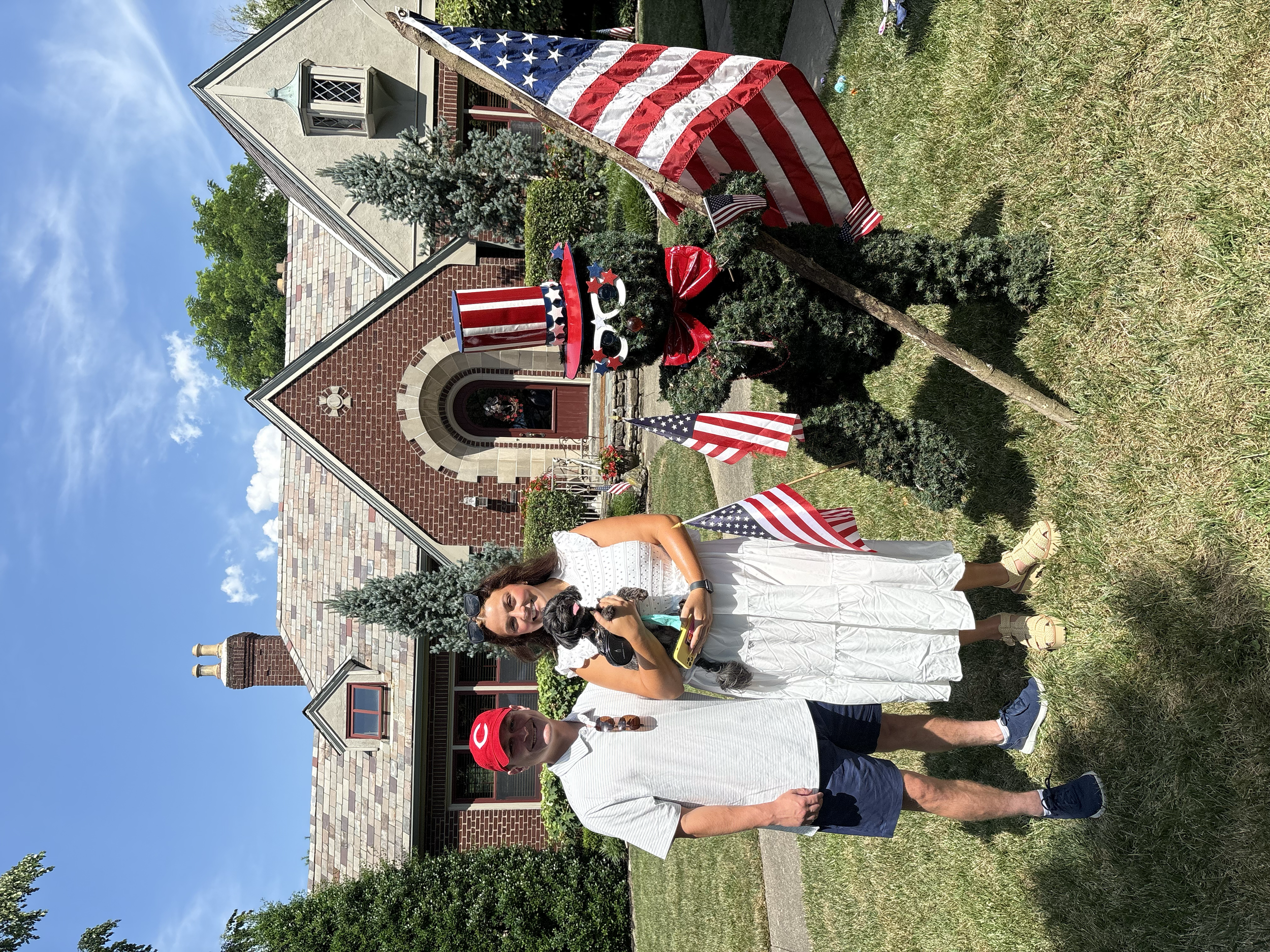 A woman and a man posing with American flags in front of a small building decorated with patriotic decorations, including a large American flag, red, white, and blue themed accessories, and a red bow, on a grassy area under a blue sky with some cloud