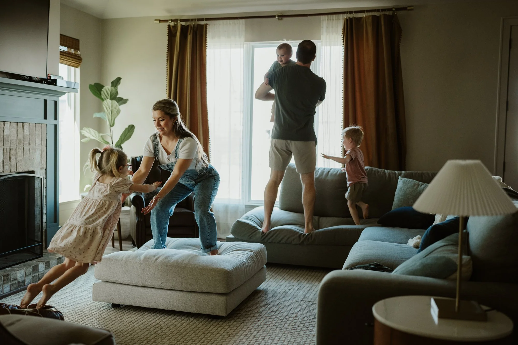 A family of five playing and jumping in a living room with sunlight coming through the windows, including a mother, father, and three children.