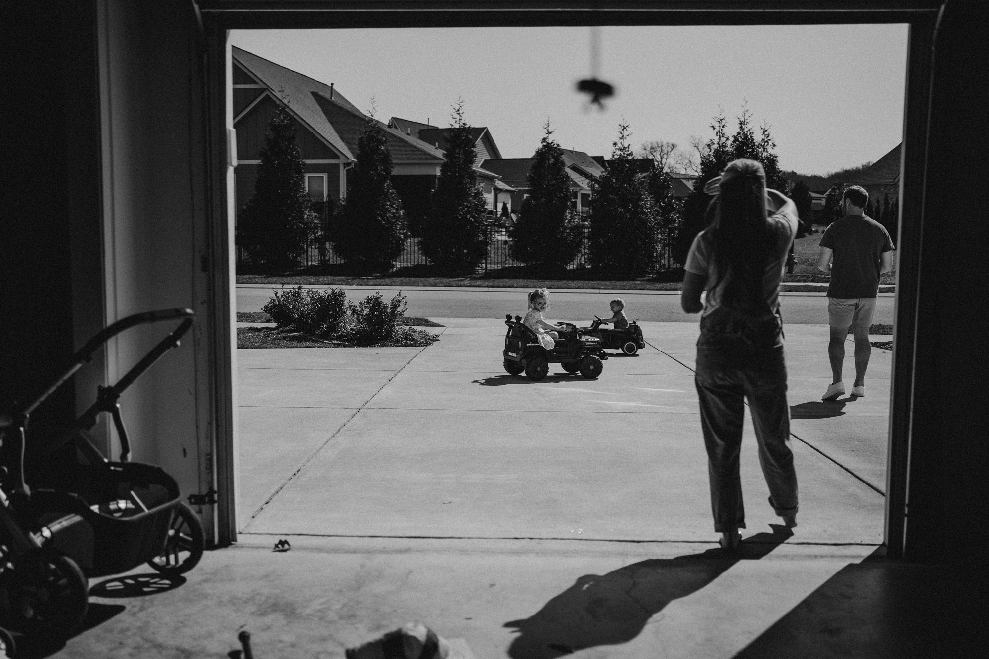 View from inside a garage looking out onto the driveway with children playing on electric toy vehicles and adults nearby on the street in a suburban neighborhood.