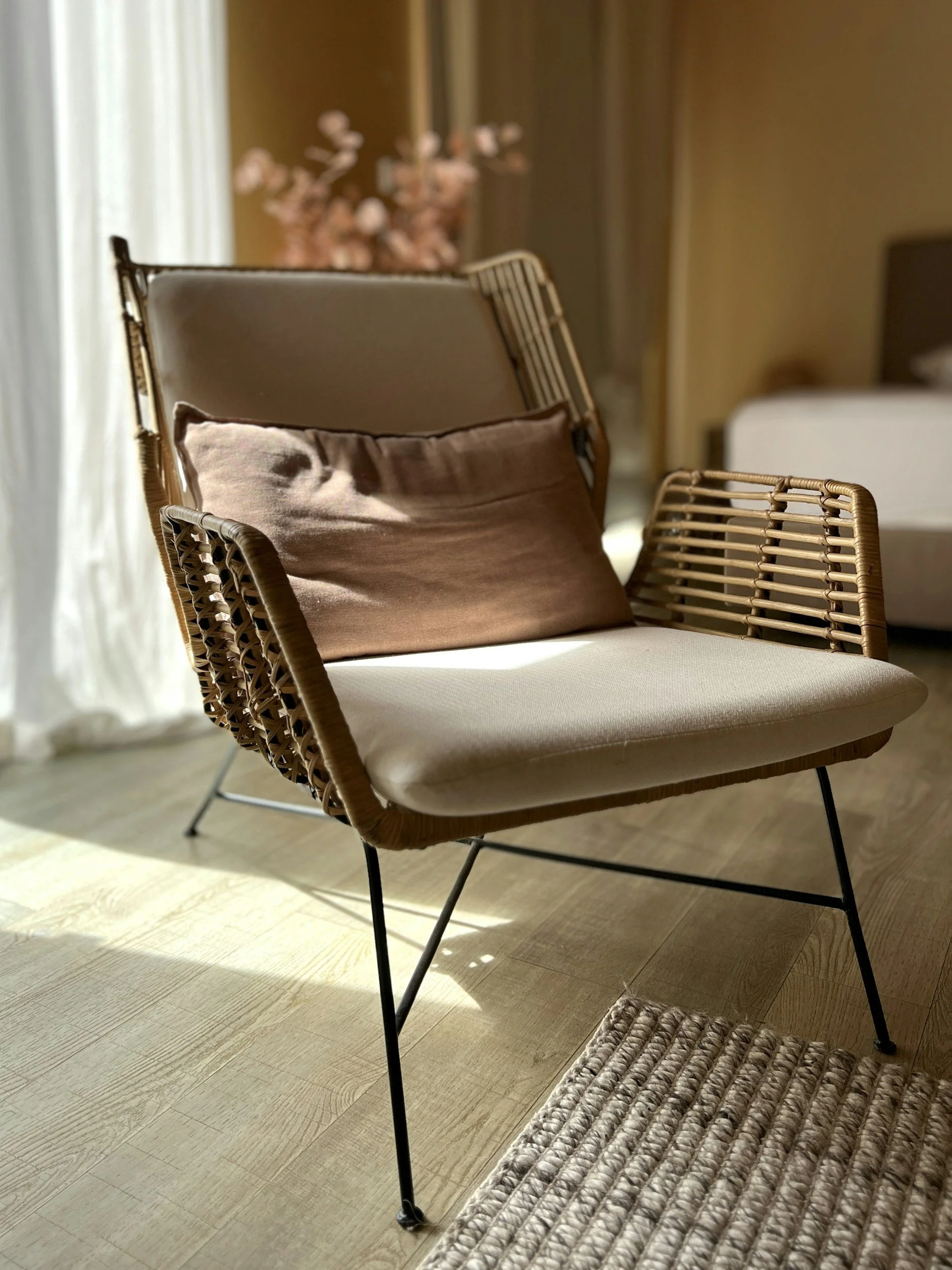 A modern armchair with a rattan frame and black metal legs, with a brown pillow placed on it, situated on a wooden floor near a beige rug, in a cozy room with sunlight coming through a window and a bed in the background.