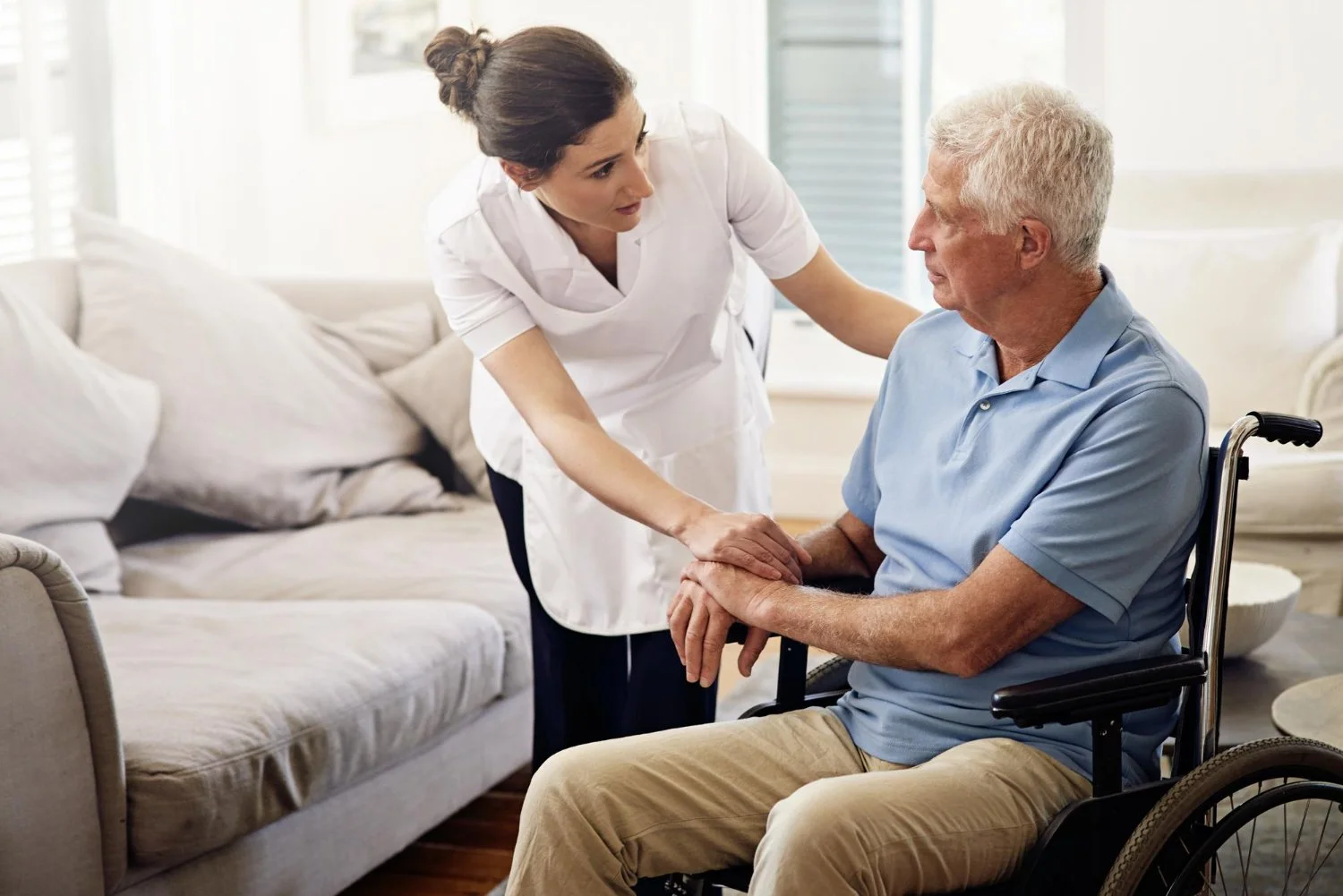 A woman caregiver gently holding hands with an elderly man in a wheelchair, inside a bright living room.