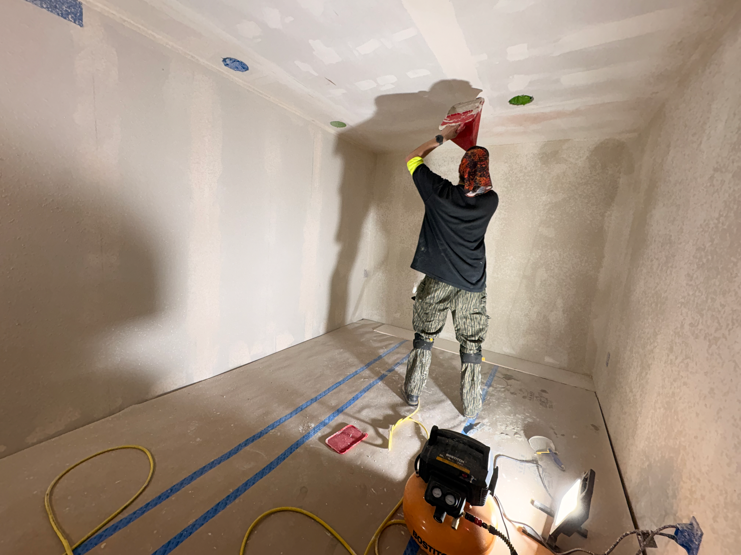 A person wearing a black shirt, patterned pants, and a head covering is painting a wall with a roller brush in a room under construction. The floor is covered with protective material, and painting tools and equipment are visible on the floor.