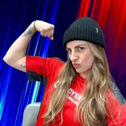 A woman with long, wavy hair smiling at the camera, wearing a black turtleneck sweater, against a plain light-colored wall.