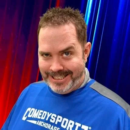 A smiling man with short dark and gray hair, a beard, and mustache, wearing a patterned button-up shirt, standing against a plain beige wall.
