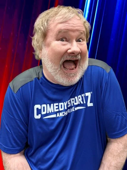 A smiling man with gray hair and beard wearing a blue T-shirt that says 'Local Tastes Better' and 'LAT 65' on a beige wall background.