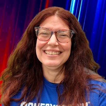 A woman with long, curly brown hair, wearing glasses and a black shirt, smiling and laughing.