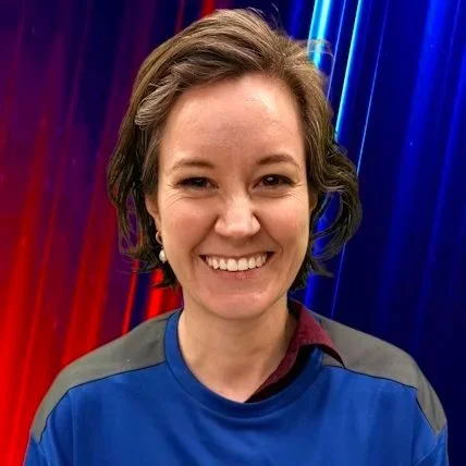 A woman with shoulder-length wavy brown hair smiling, wearing a black, white, and orange patterned sweater and a gold necklace, against a plain brown background.