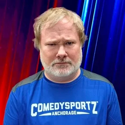 A smiling man with gray hair and beard wearing a blue T-shirt that says 'Local Tastes Better' and 'LAT 65' on a beige wall background.
