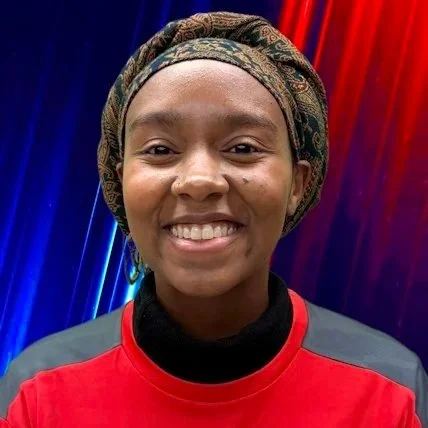 Smiling young woman with natural hair in a puff, wearing a checkered shirt over a blue top, standing against a beige wall.