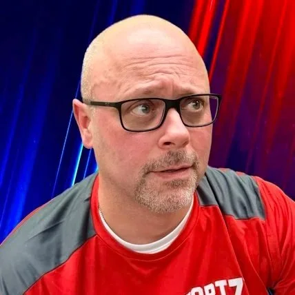 Smiling man with a bald head, beard, wearing a black vest over a checkered shirt, standing against a plain beige wall.