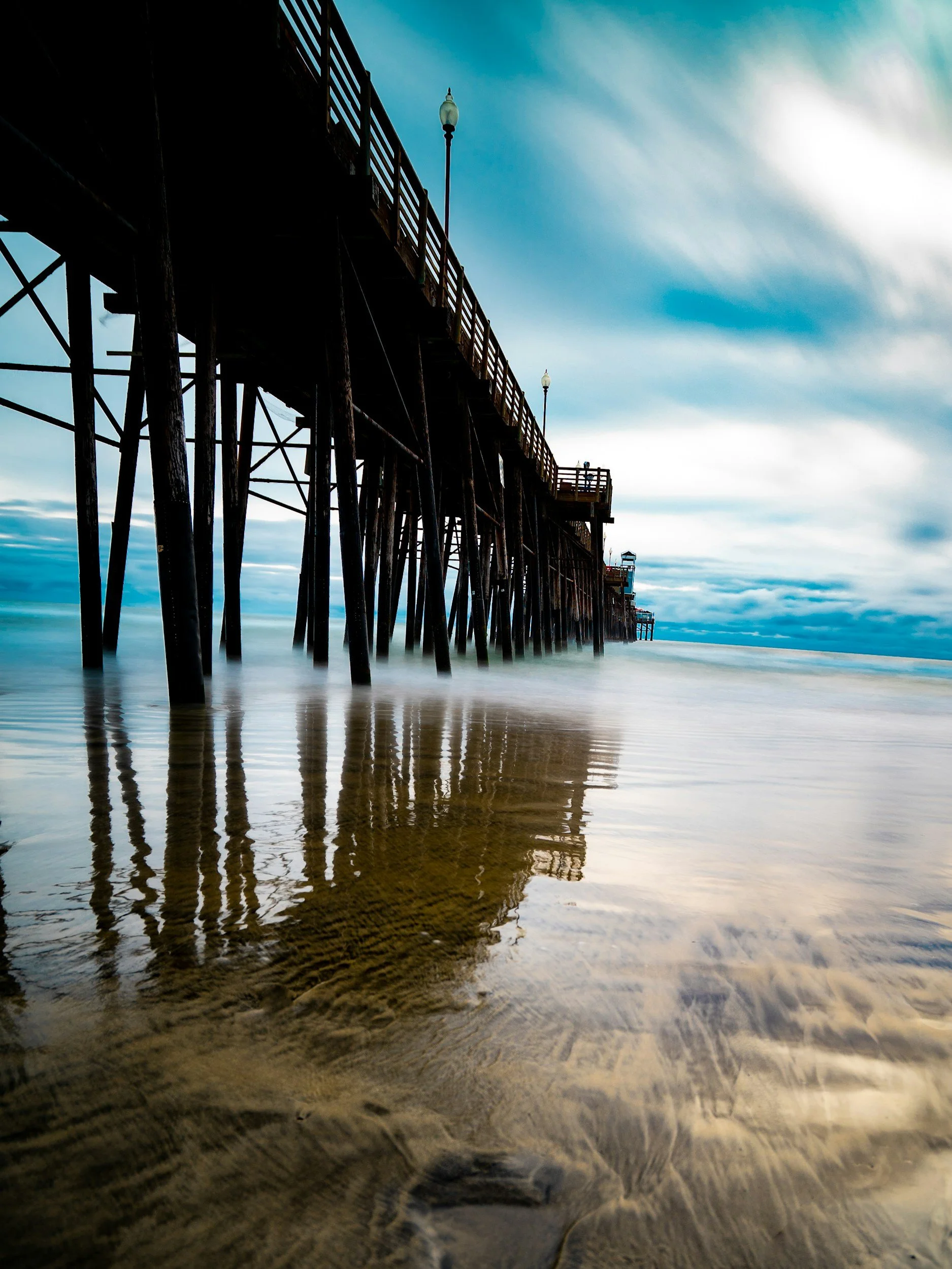 View of a wooden pier extending into the ocean, with water underneath and a cloudy sky overhead.