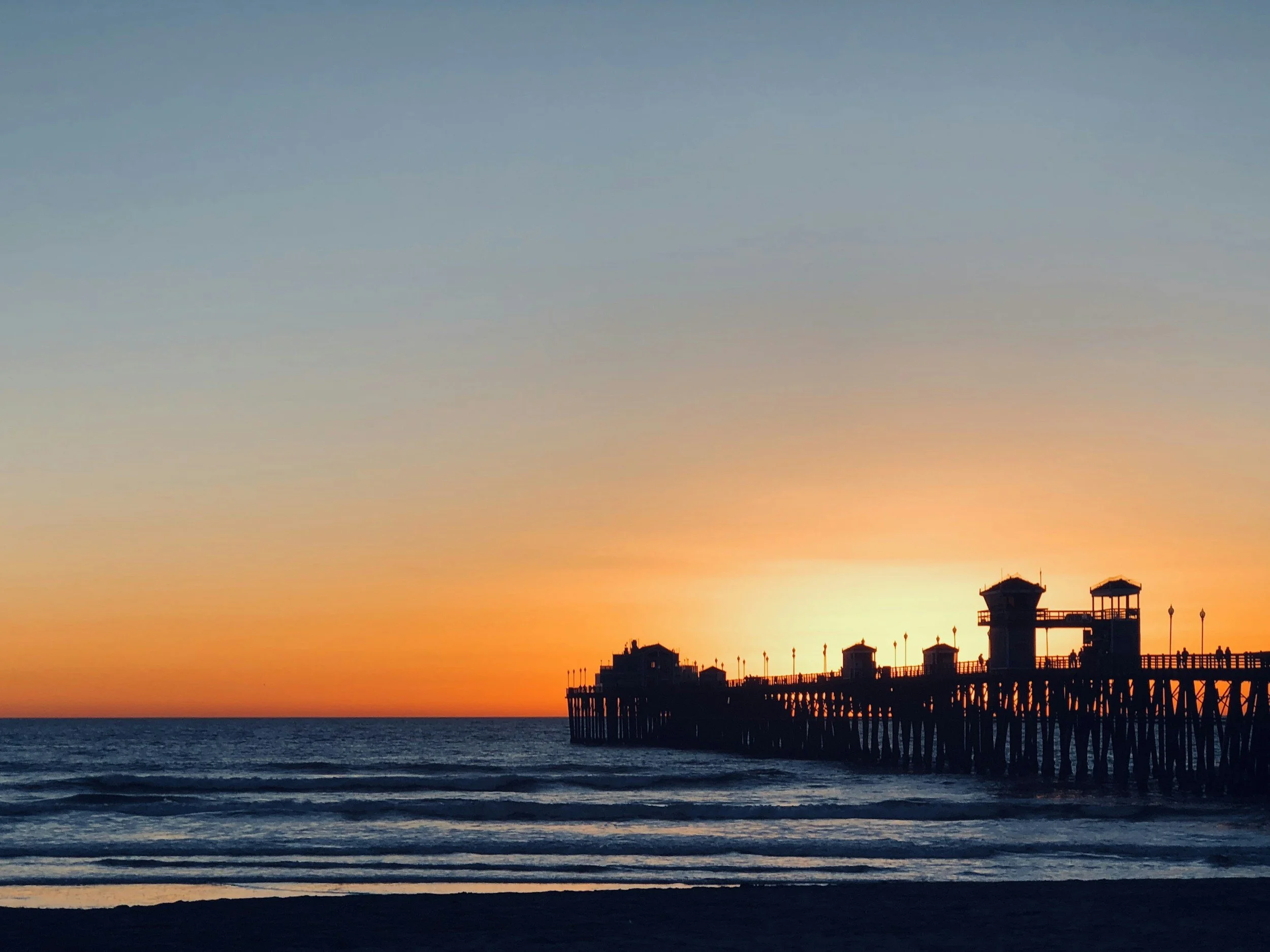 Sunset at the ocean with a pier extending into the water, silhouetted structures on the pier, and a clear sky transitioning from orange near the horizon to blue higher up.
