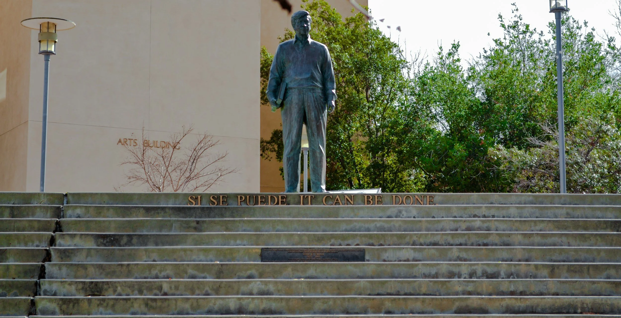 CSUSM Statue of a man, Cesar Chavez, holding books, standing on steps with the phrase 'SI SE PUEDE IT CAN BE DONE' inscribed below. Background includes trees, a building with the words 'ARTS BUILDING', and street lamps in San Marcos, Ca.