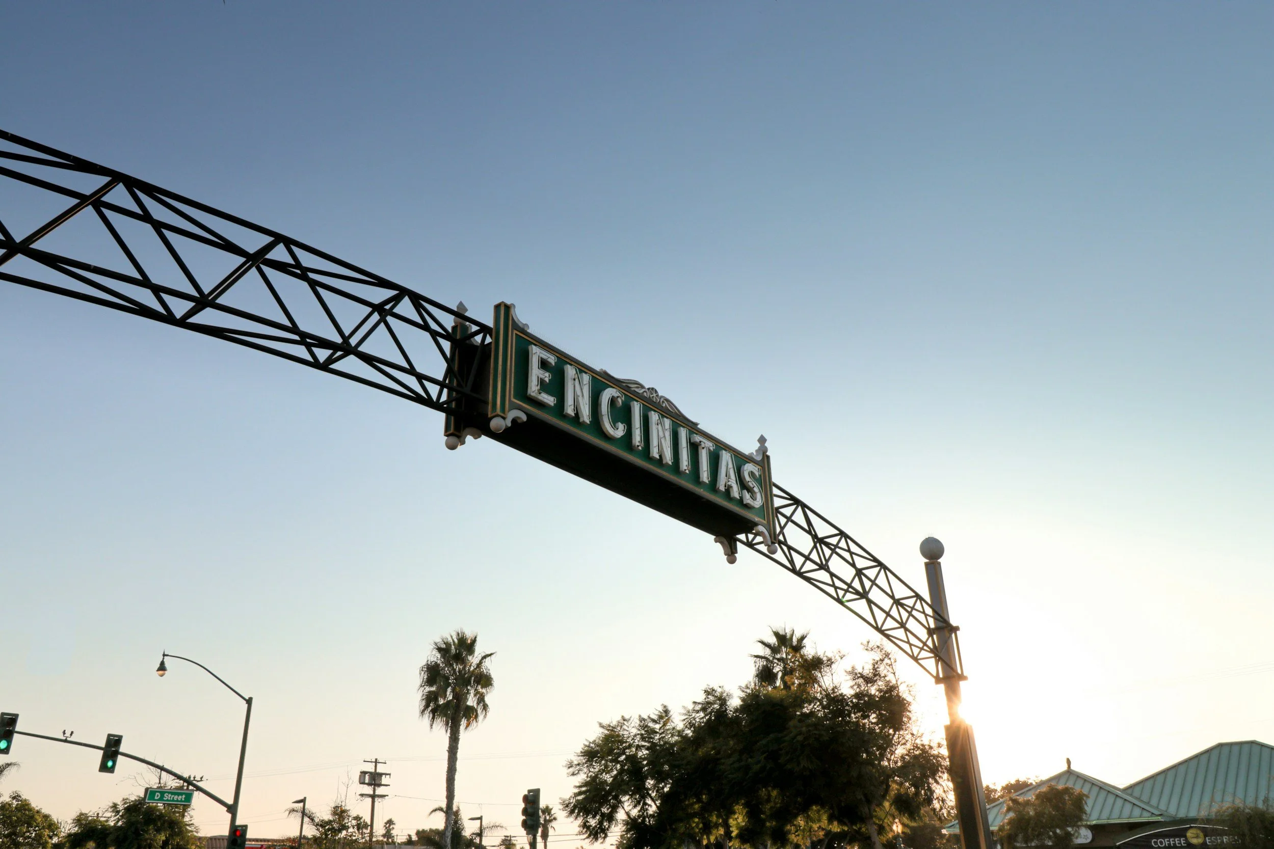 View of Encinitas street scene at sunset with the large green sign reading 'Encinitas' hanging from a metal frame, palm trees, traffic lights, and a building with a metal roof in the background.