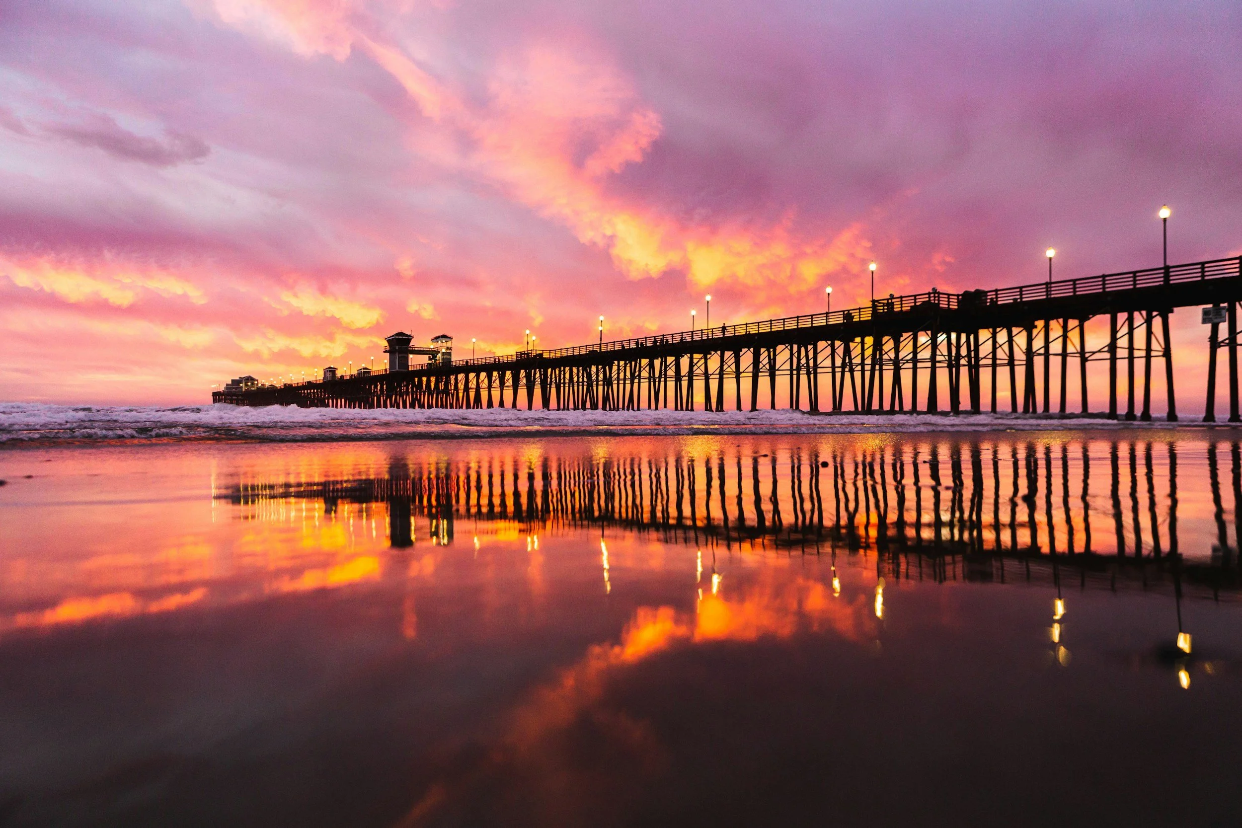 Sunset at the beach with a pier extending into the ocean, colorful sky with pink and orange clouds, reflection of the pier and sky on the wet sand.