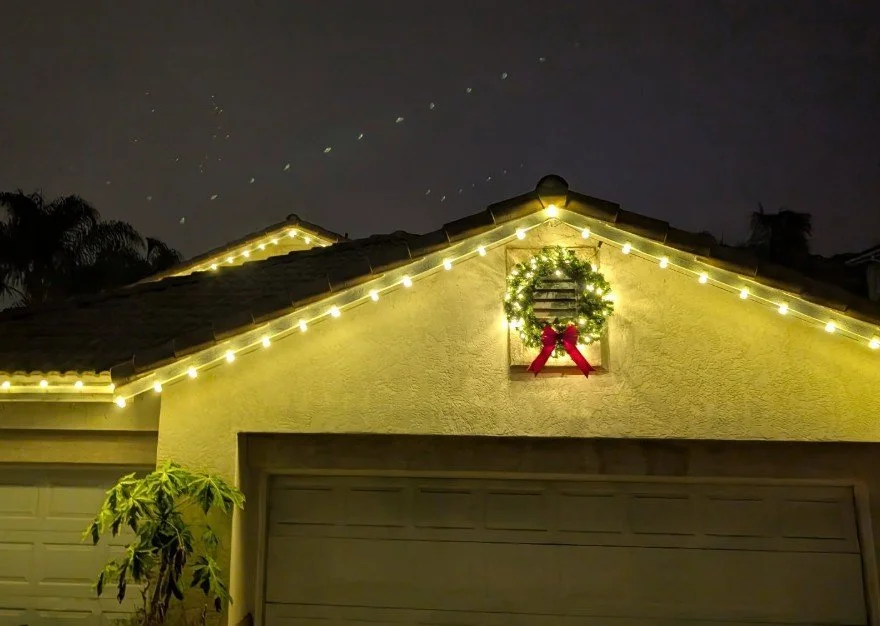 House with Christmas lights along the roof line, a Christmas wreath with a red bow in a small window, and a starry night sky overhead.