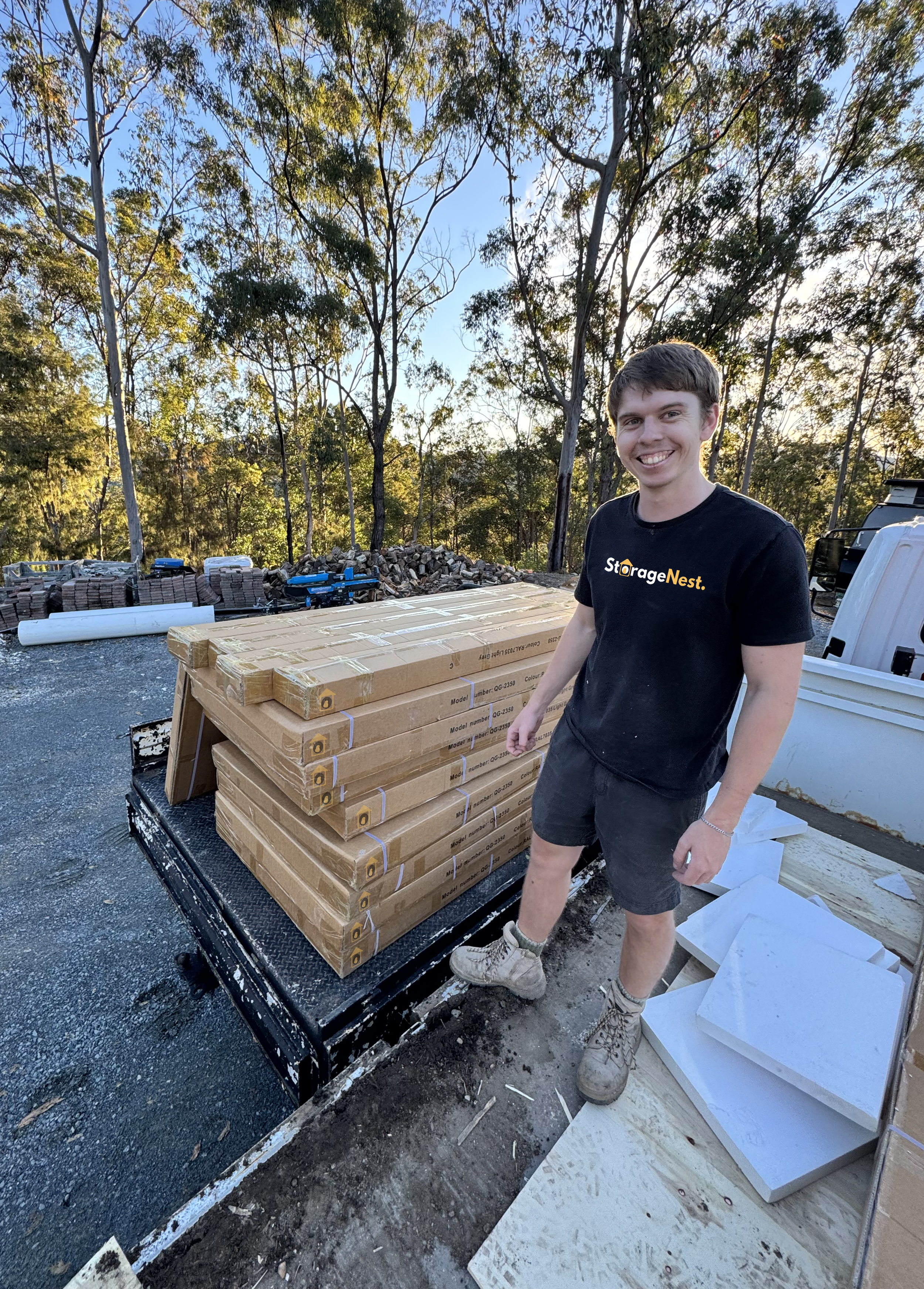 A smiling young man in work clothes standing next to a load of boxed tiles on a flatbed trailer in an outdoor construction or landscaping site surrounded by trees. Unloading stock of over bonnet storage boxes.