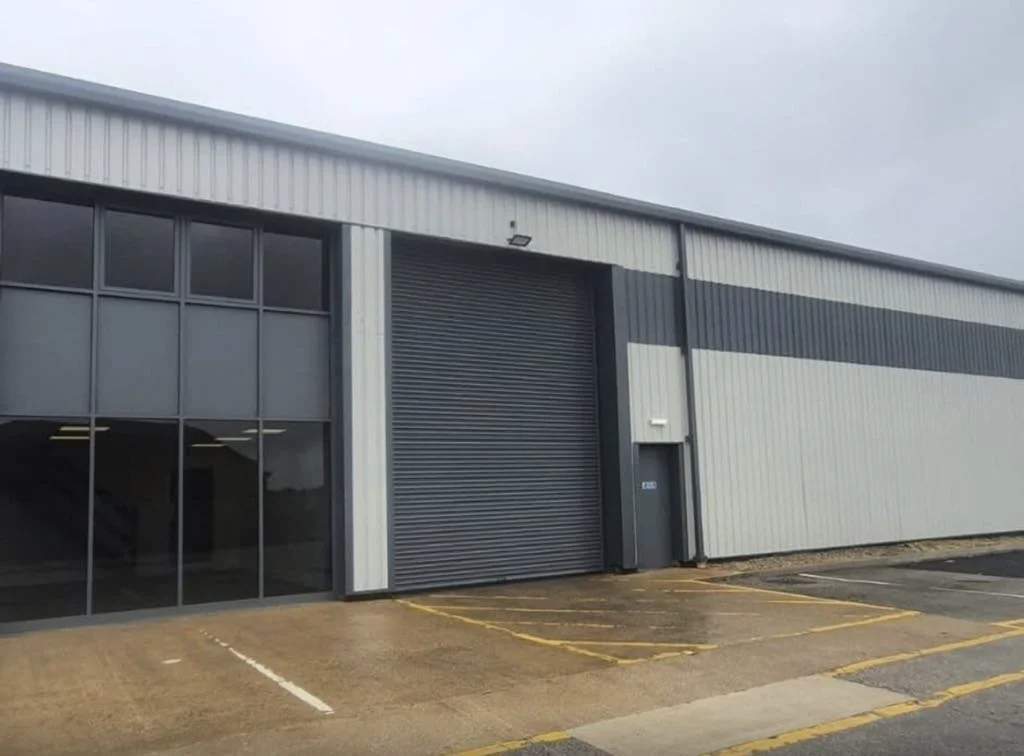 Exterior of a commercial warehouse with large glass windows, a roll-up door, and a small side door, with parking spaces in front under an overcast sky.