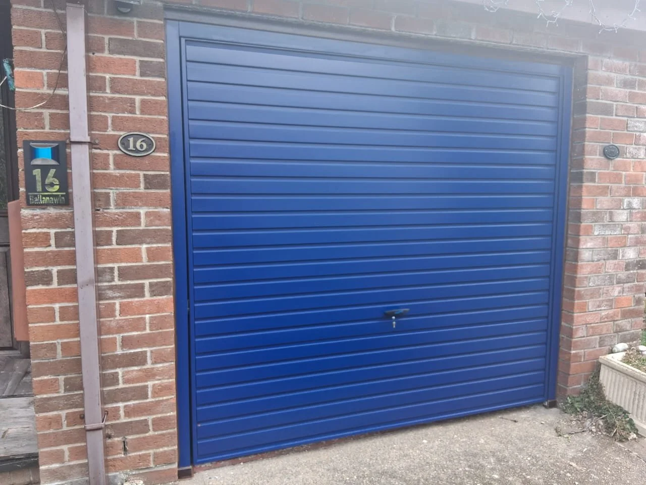 A blue metal garage door on a brick house with house number 16 and a mailbox nearby.