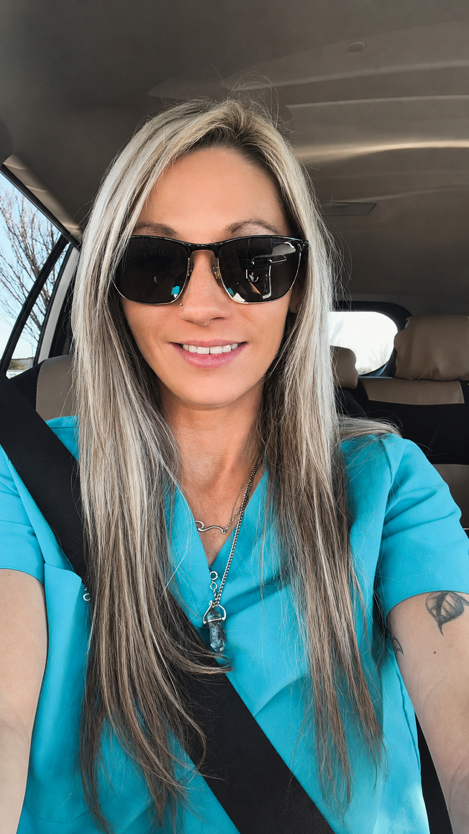 A woman with long blonde hair wearing sunglasses and a blue medical uniform taking a selfie inside a vehicle.