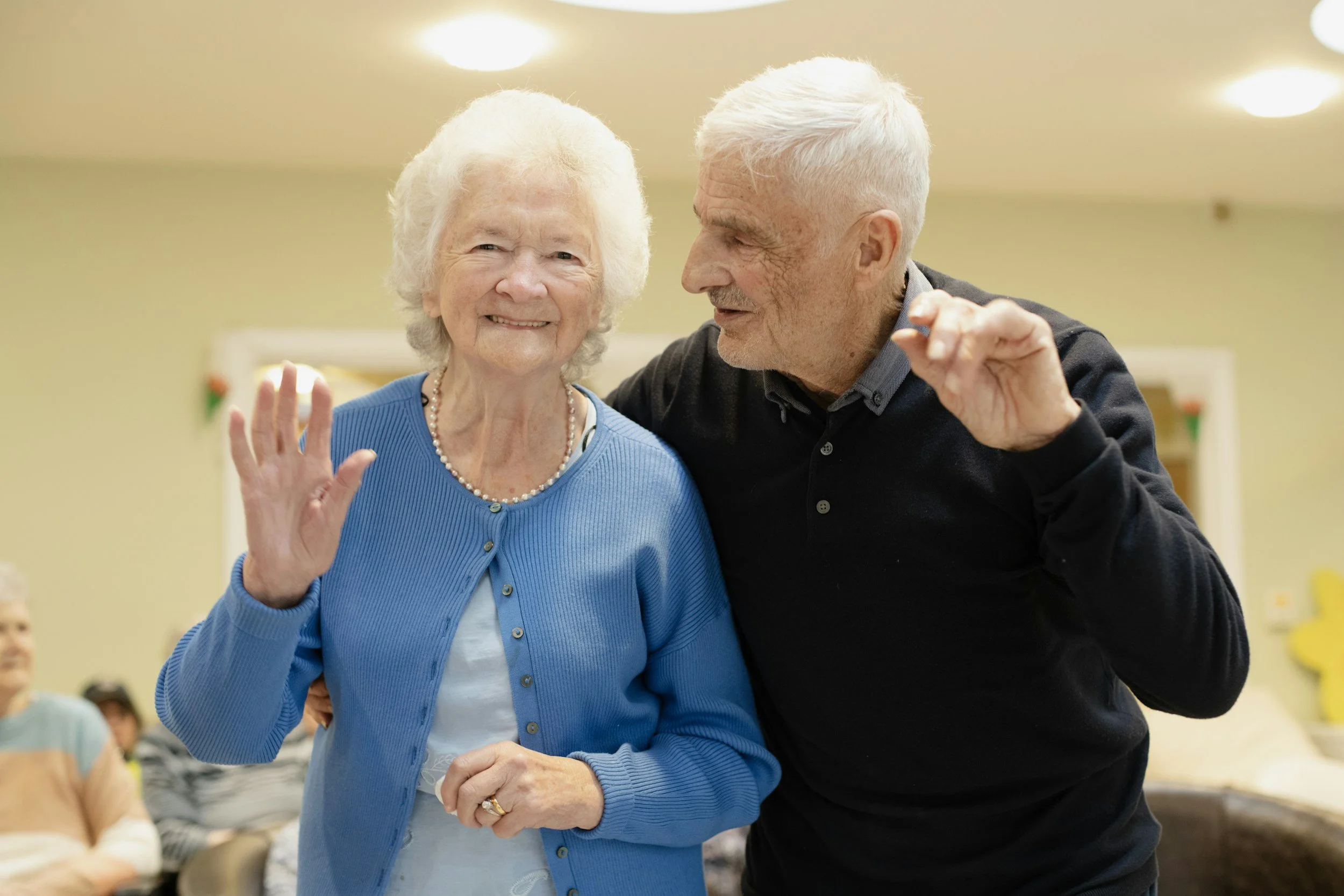 An elderly woman wearing a blue cardigan and a pearl necklace smiling and waving, standing beside an elderly man with white hair, who is leaning in close and speaking to her, at a social gathering or event.