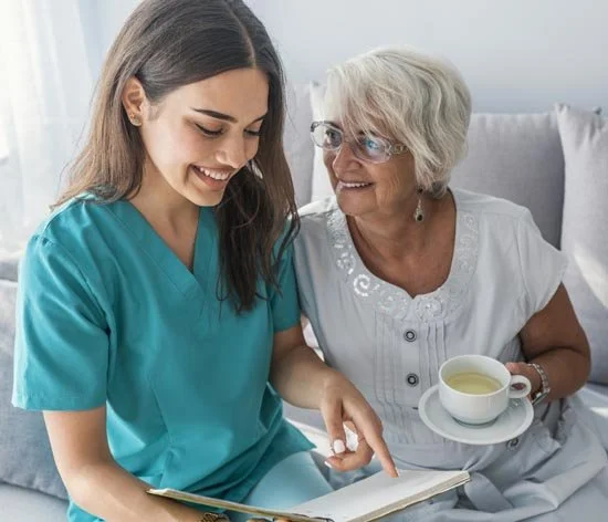 A young woman in medical scrubs reading a book to an elderly woman who is holding a cup of tea, both smiling.