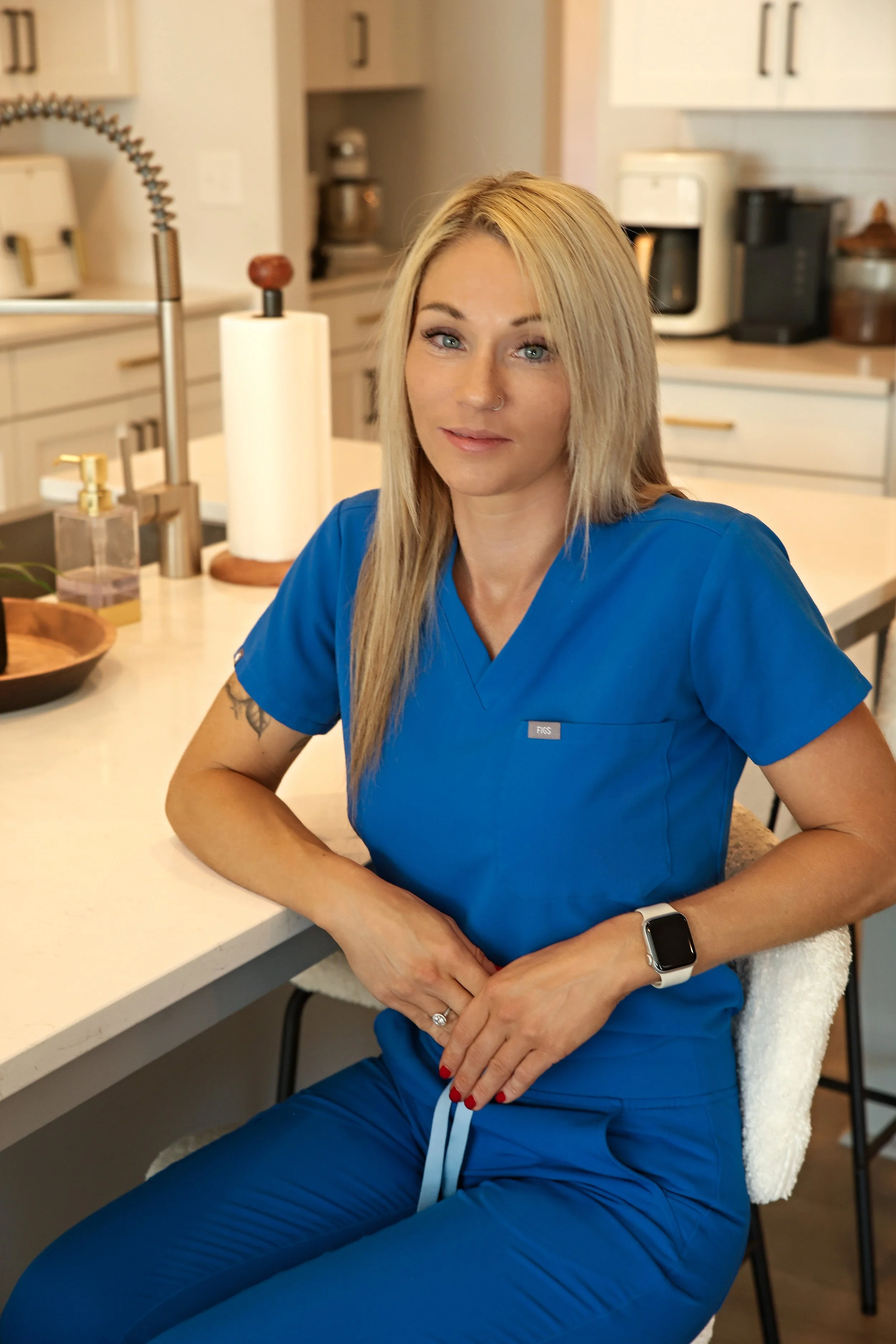 A woman in blue medical scrubs sitting at a kitchen counter with her arms crossed, wearing a smartwatch, with kitchen appliances in the background.