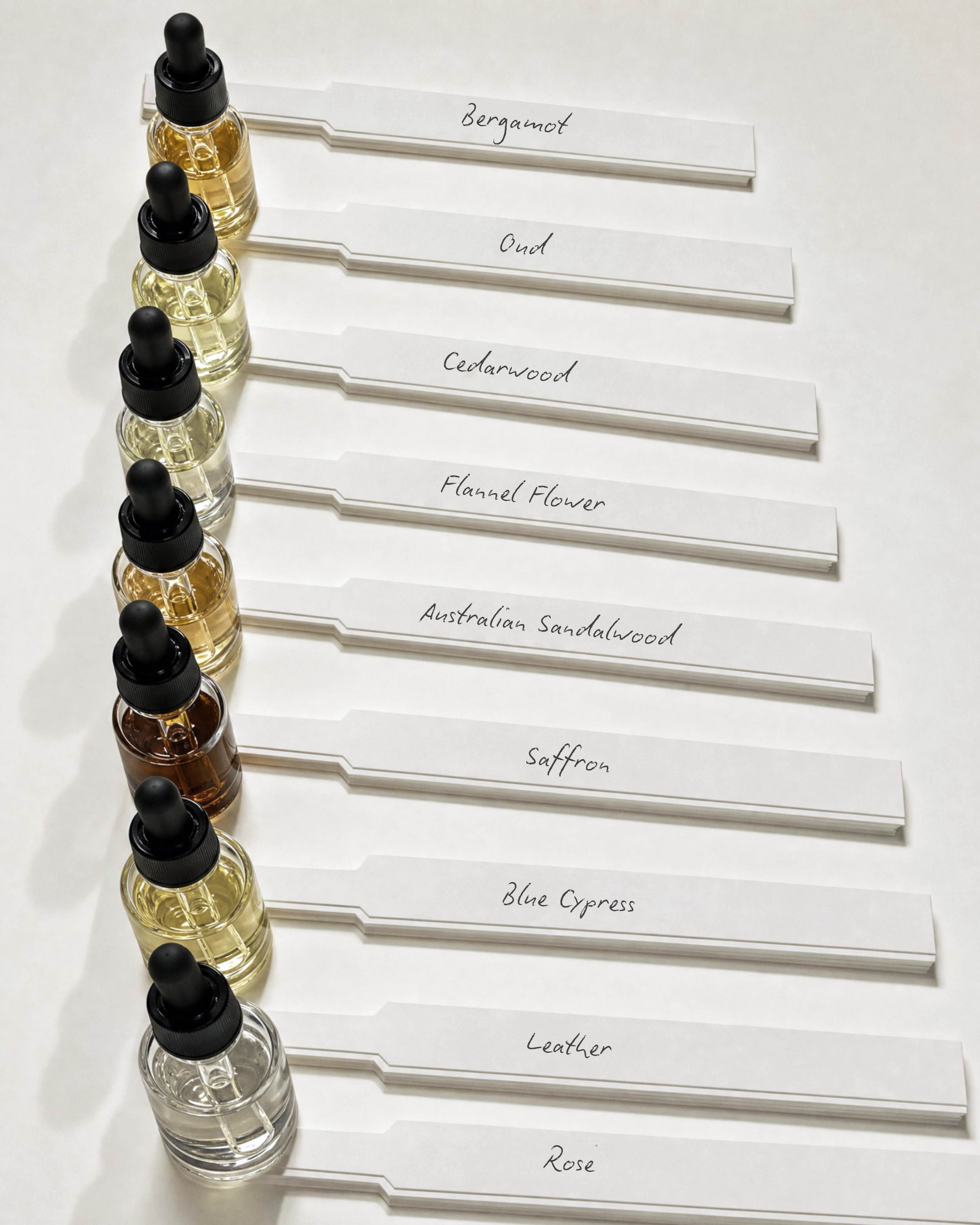Six small glass bottles with black droppers, each filled with a different light-colored liquid, arranged in a vertical line. Each bottle has a white paper label with handwritten names of fragrances: Bergamot, Oud, Cedarwood, Flannel Flower, Australian Sandalwood, and Rose.