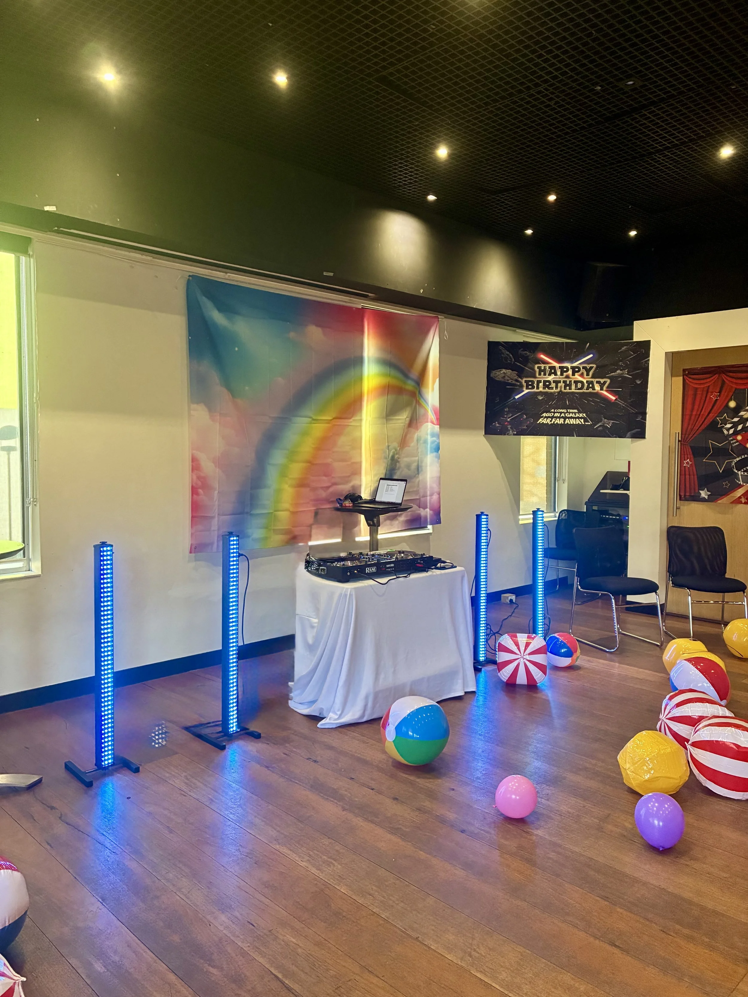 A decorated party room with beach balls, helium balloons, a rainbow backdrop, a DJ setup with blue standing LED lights, and a 'Happy Birthday' sign on the wall.