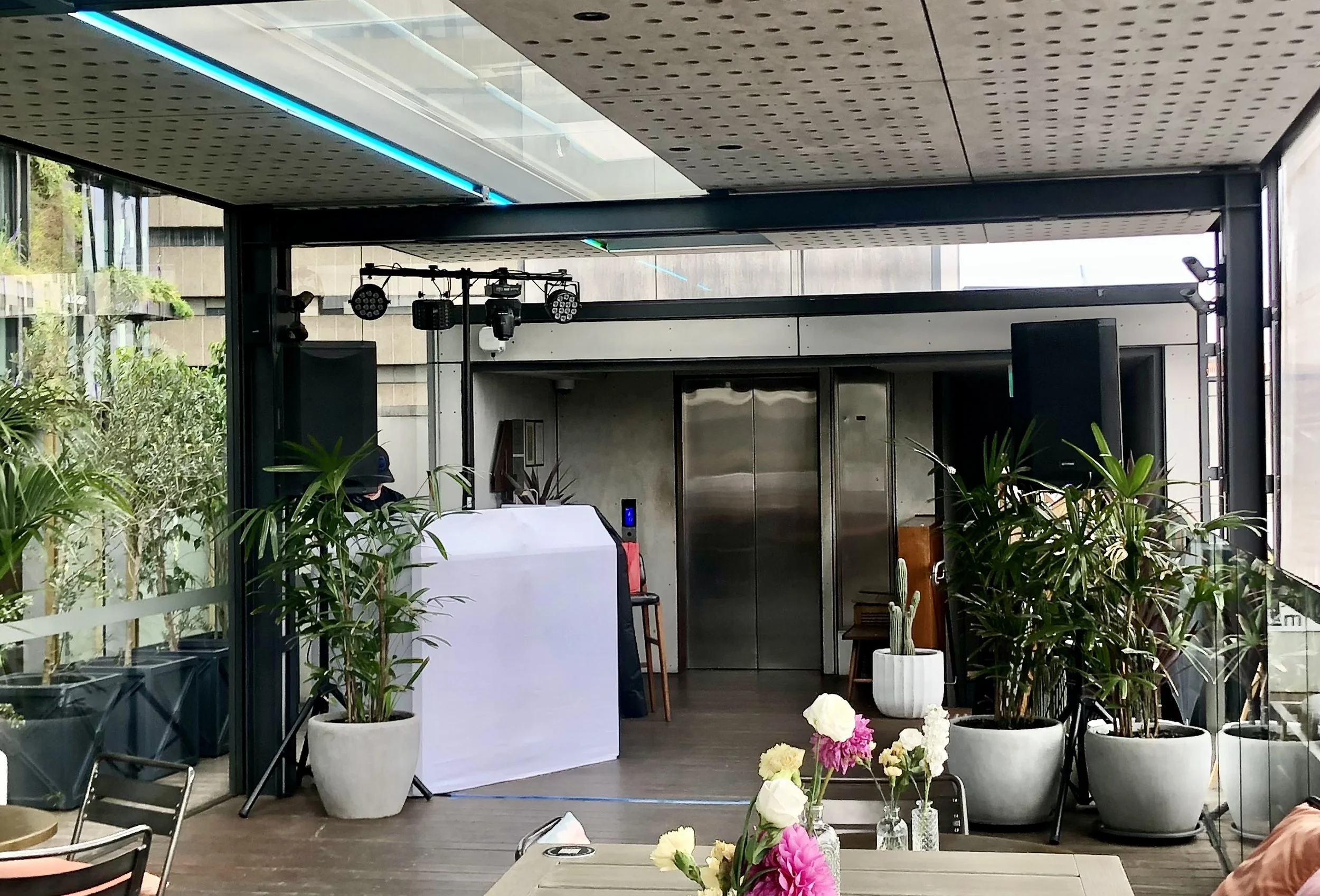Indoor space with potted plants, flowers, and a DJ booth with lighting equipment, large windows, and modern decor.