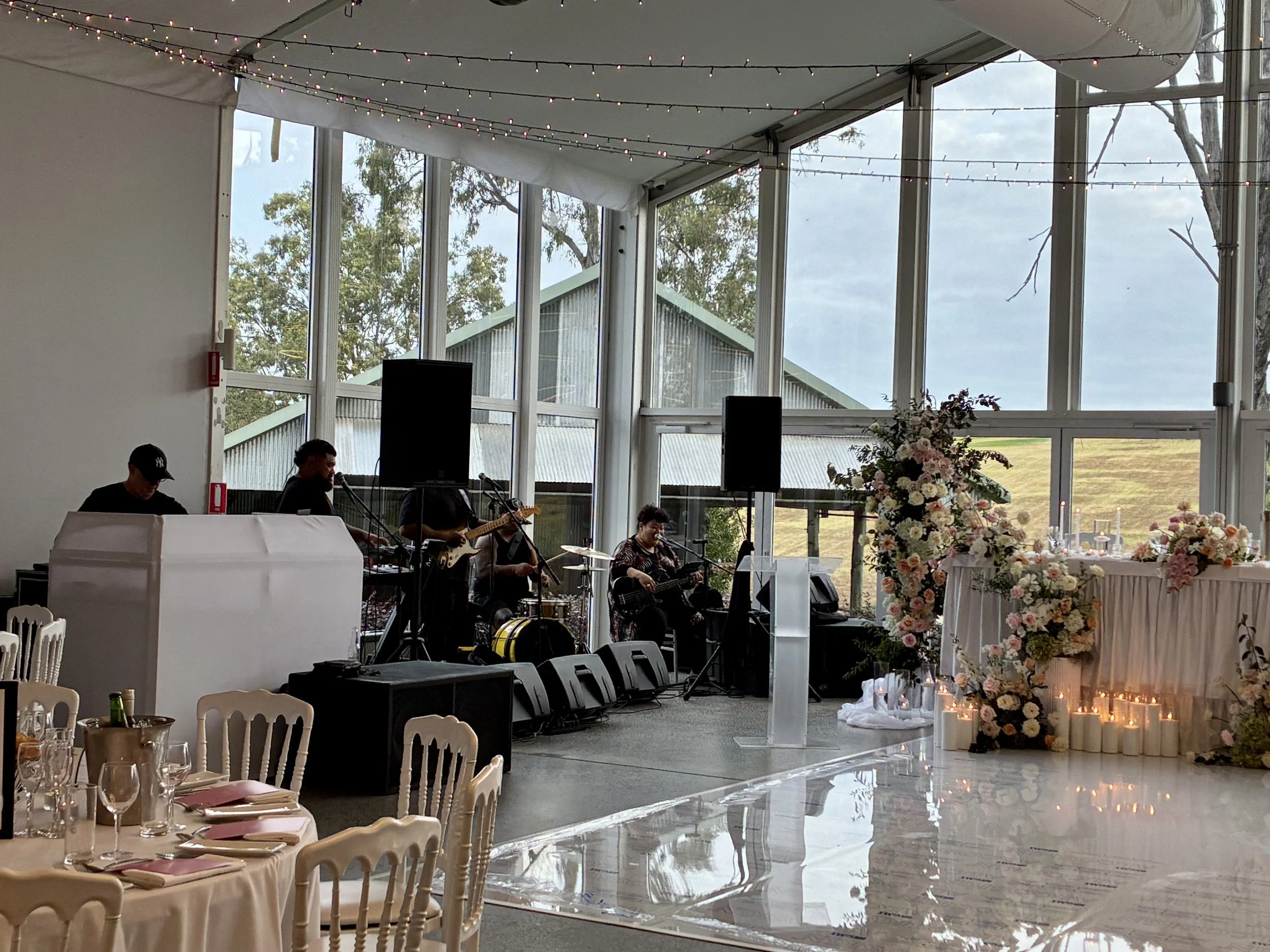 Indoor wedding reception with a deejay and a band playing music, decorated with string lights and floral arrangements, large windows showing trees and a cloudy sky outside.