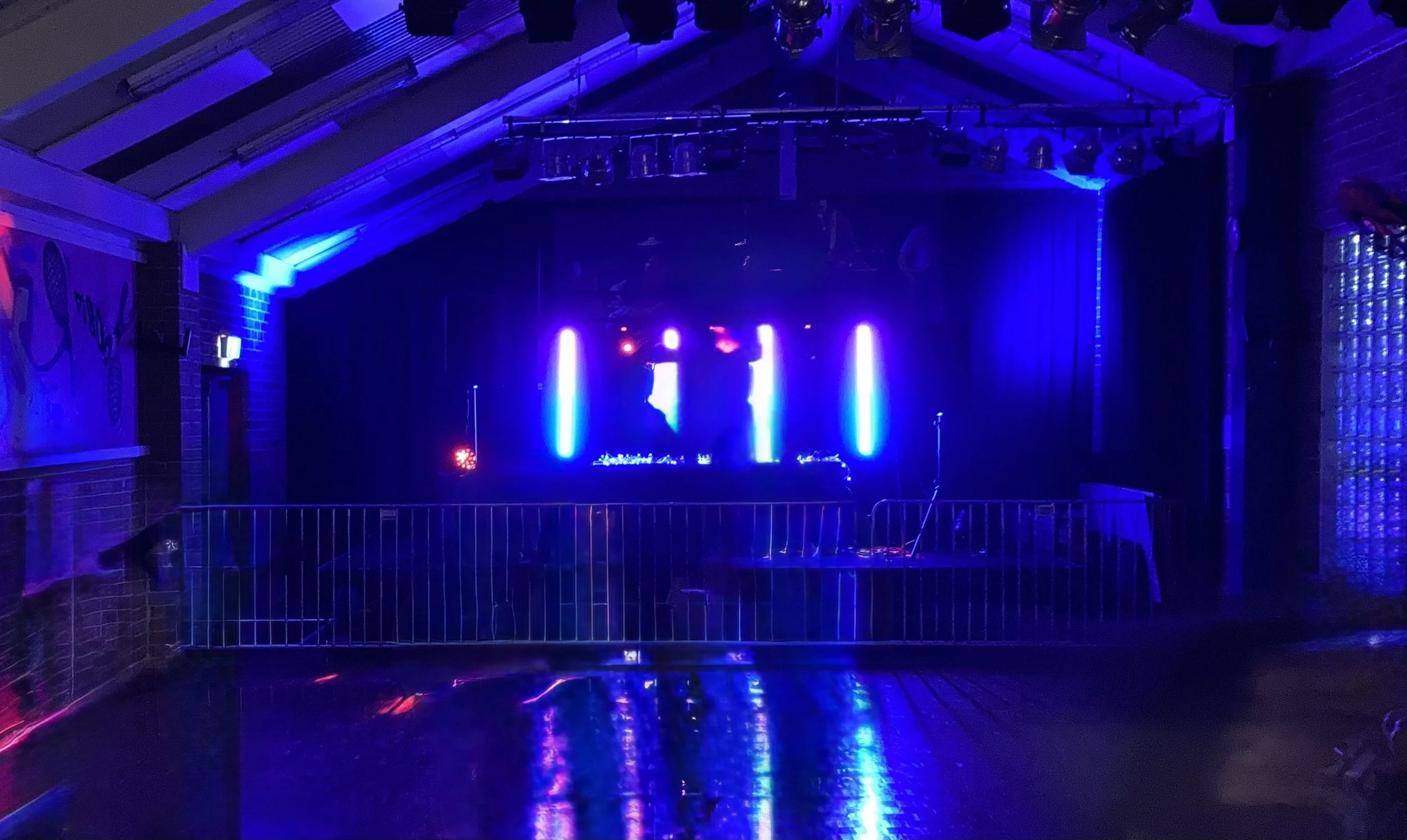 A dark room with a stage illuminated by blue and purple lights, set up for a performance or DJ event, with a black curtain backdrop and lighting fixtures overhead.