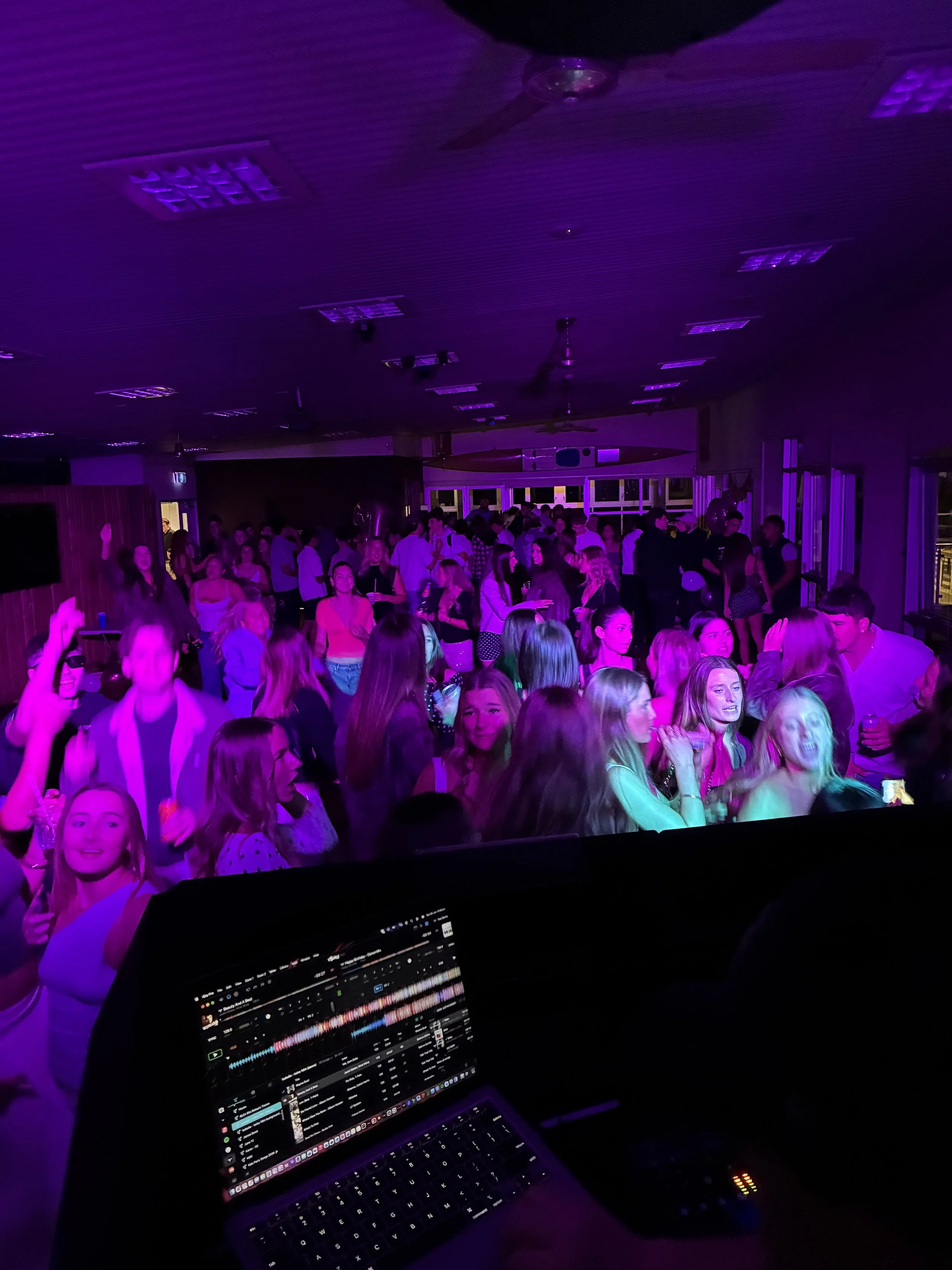 A DJ's laptop displaying music tracks and waveforms, with a crowded dance floor of people dancing and socializing illuminated by purple lighting in a nightclub or party setting.