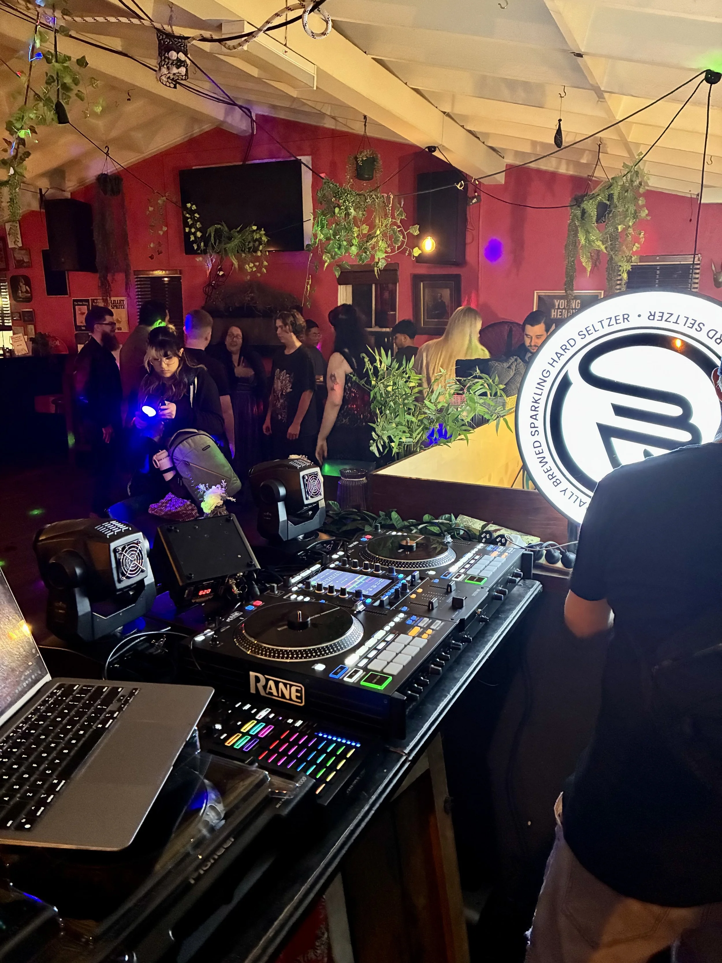 A DJ setup with a laptop, DJ controllers, and audio equipment in a dimly lit club with people dancing and socializing, adorned with hanging plants and colorful lighting.