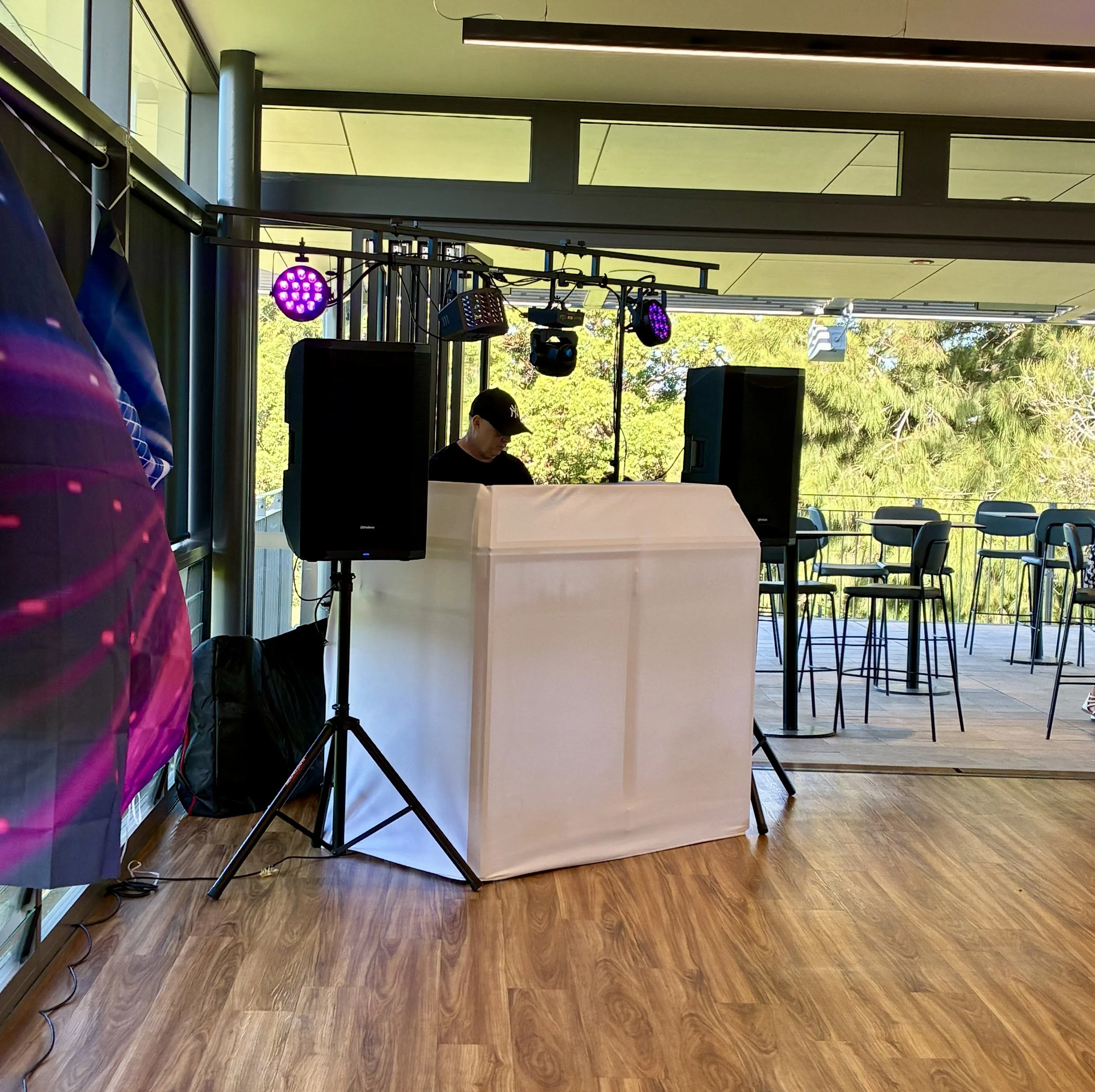 Indoor scene with colorful stage lighting, a DJ or musician wearing a black cap, a large white backdrop, chairs outside near a balcony, and large windows showing greenery outside.