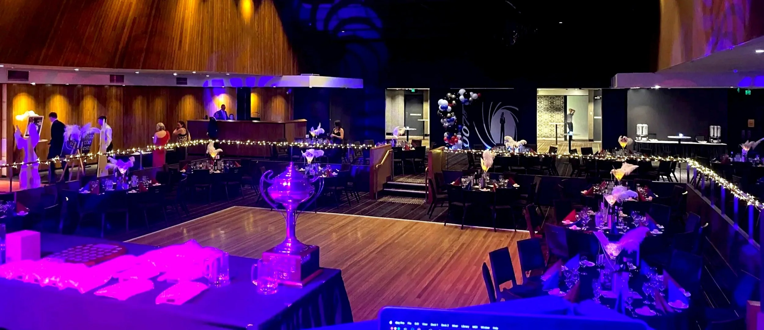 Interior of a decorated event space with tables set for a celebration, fairy lights, balloon arrangements, a bar area, and a dance floor, illuminated with purple and yellow lighting.